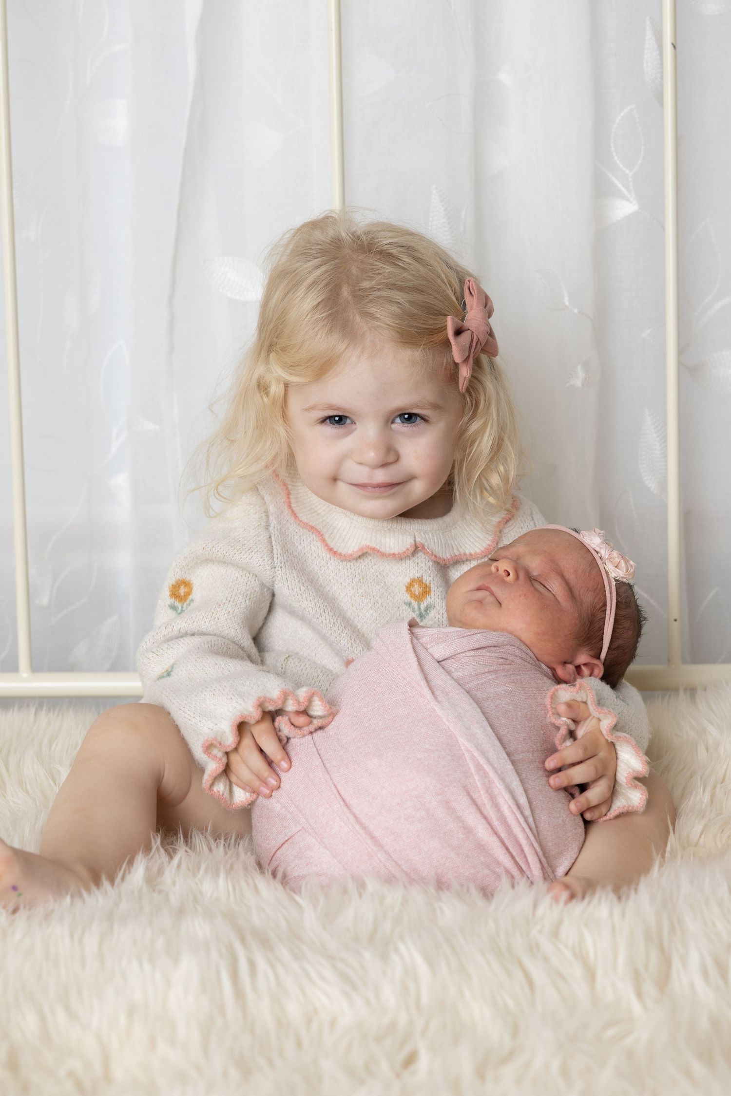 Smiling blonde toddler girl holding a sleeping newborn baby wrapped in pink on a white fluffy rug.