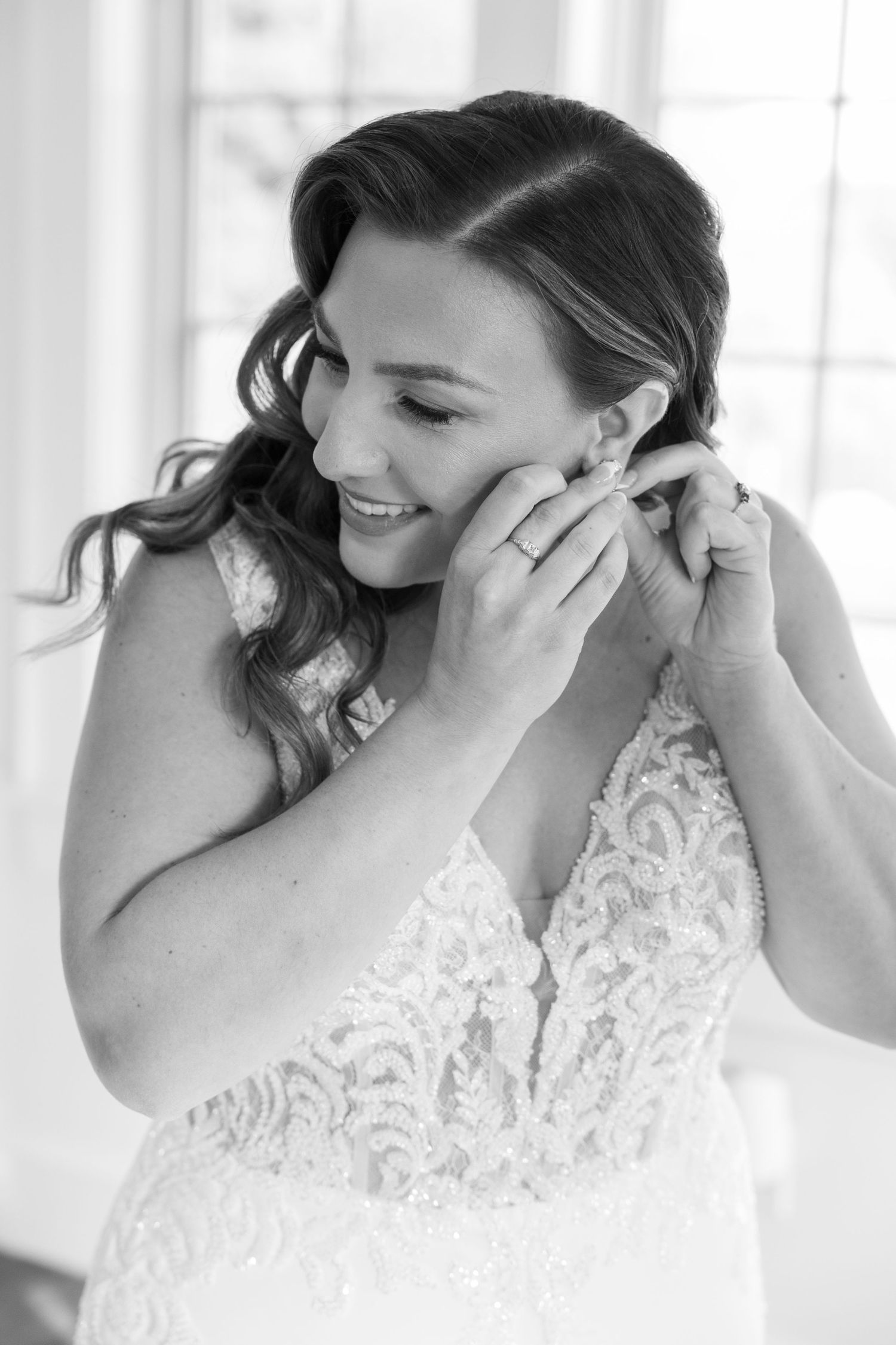 Smiling bride in lace wedding gown putting on earrings, black and white bridal portrait.