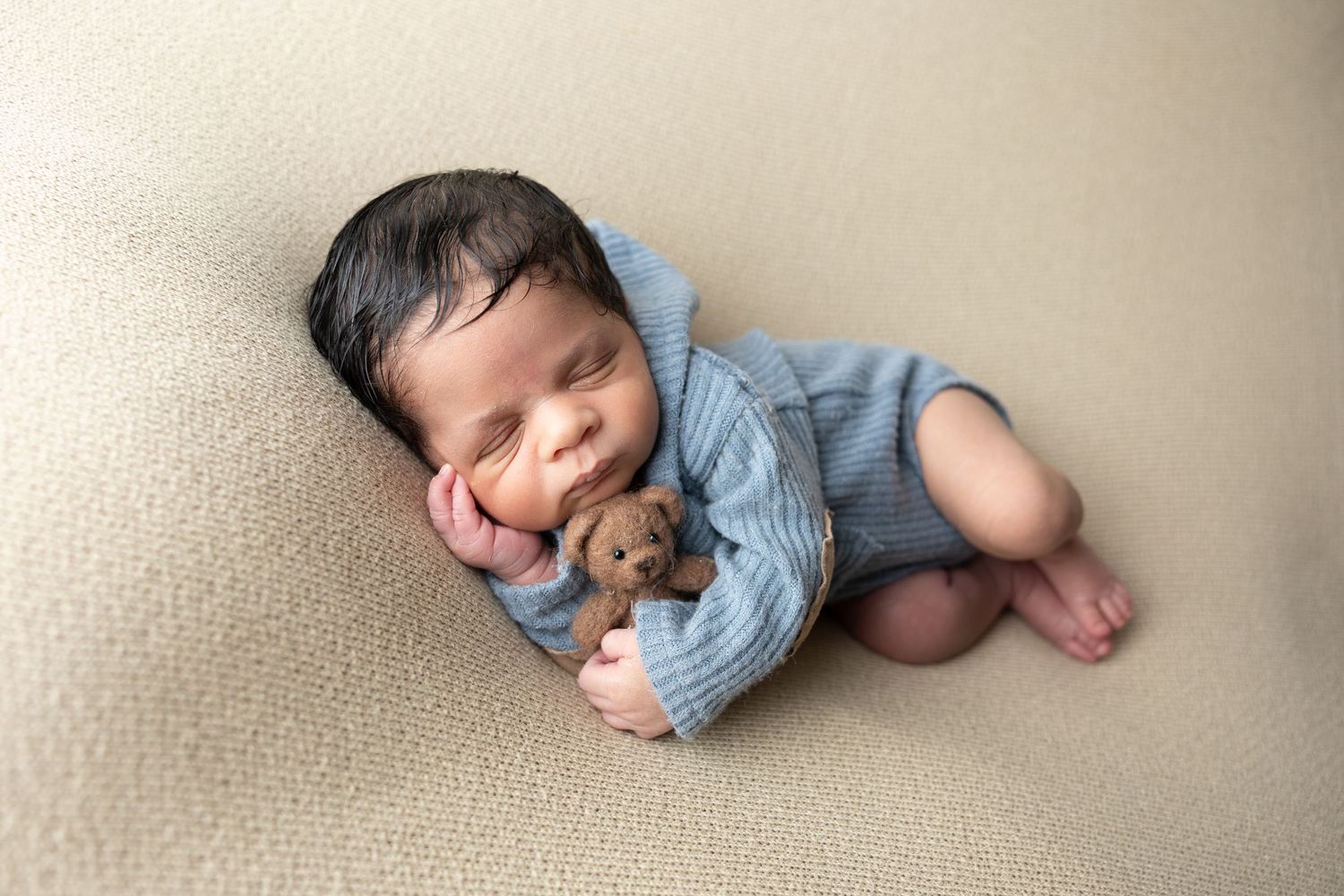 Newborn baby sleeping peacefully in blue knit romper, cuddling a tiny teddy bear on a cream blanket.
