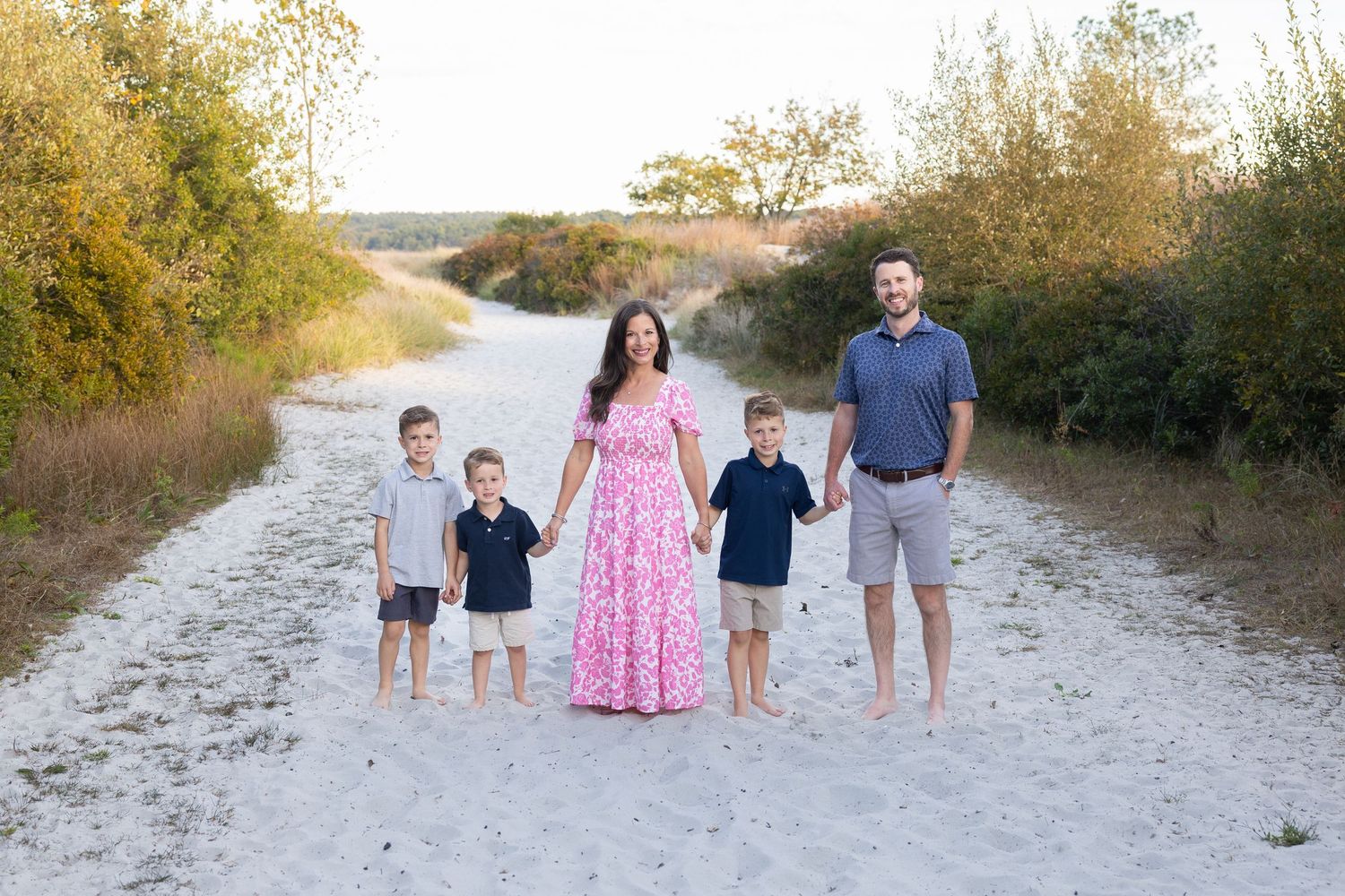 Family of five posing on sandy path surrounded by greenery, mother in pink floral dress, father in gray shirt, three young boys.
