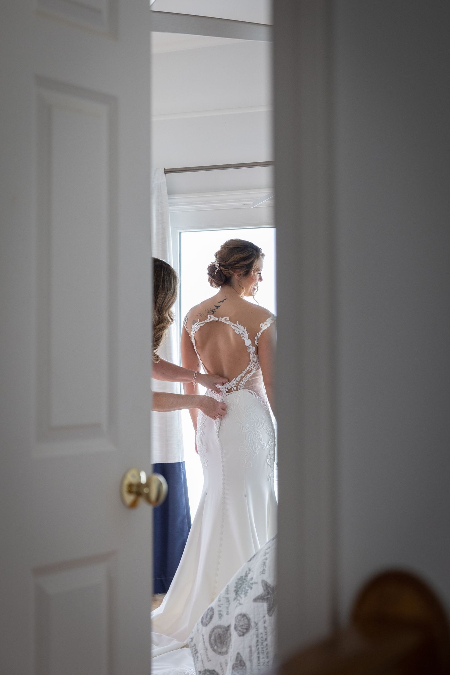 Bride getting her lace back wedding dress fastened by bridesmaid in a bright room before the ceremony.