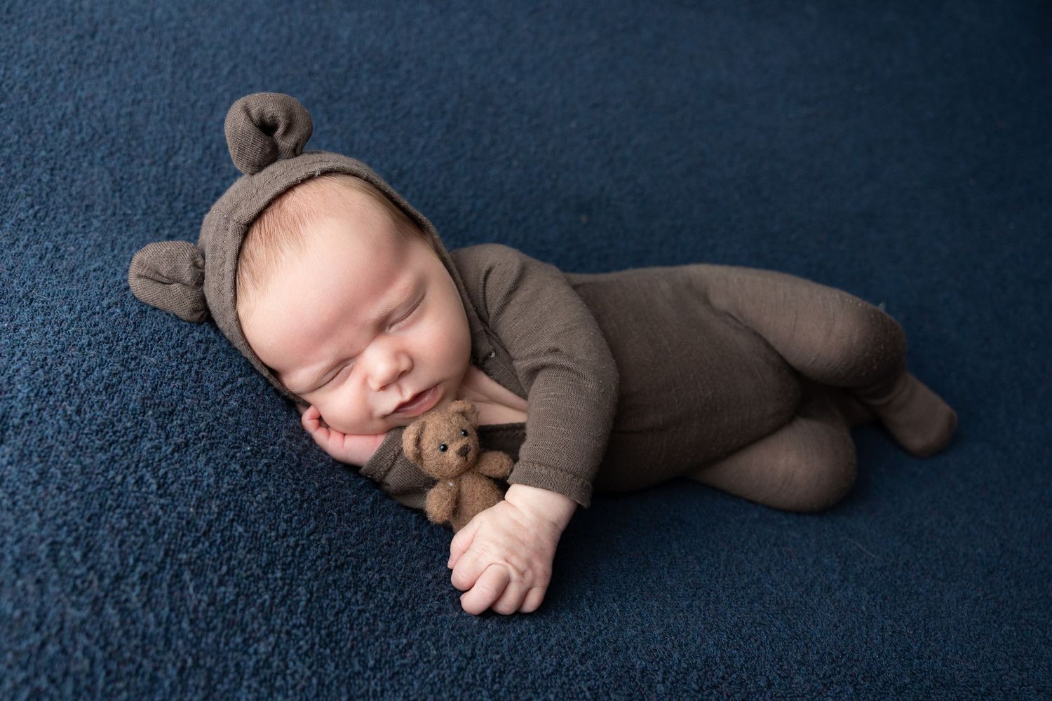Sleeping newborn baby in brown bear outfit holding tiny teddy bear on blue carpet background.