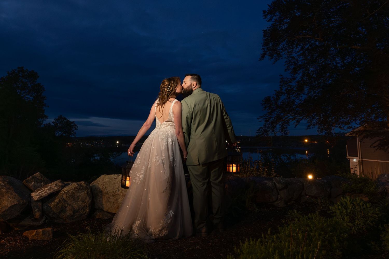 Bride and groom sharing a romantic kiss at dusk, overlooking a serene waterfront with glowing lanterns.