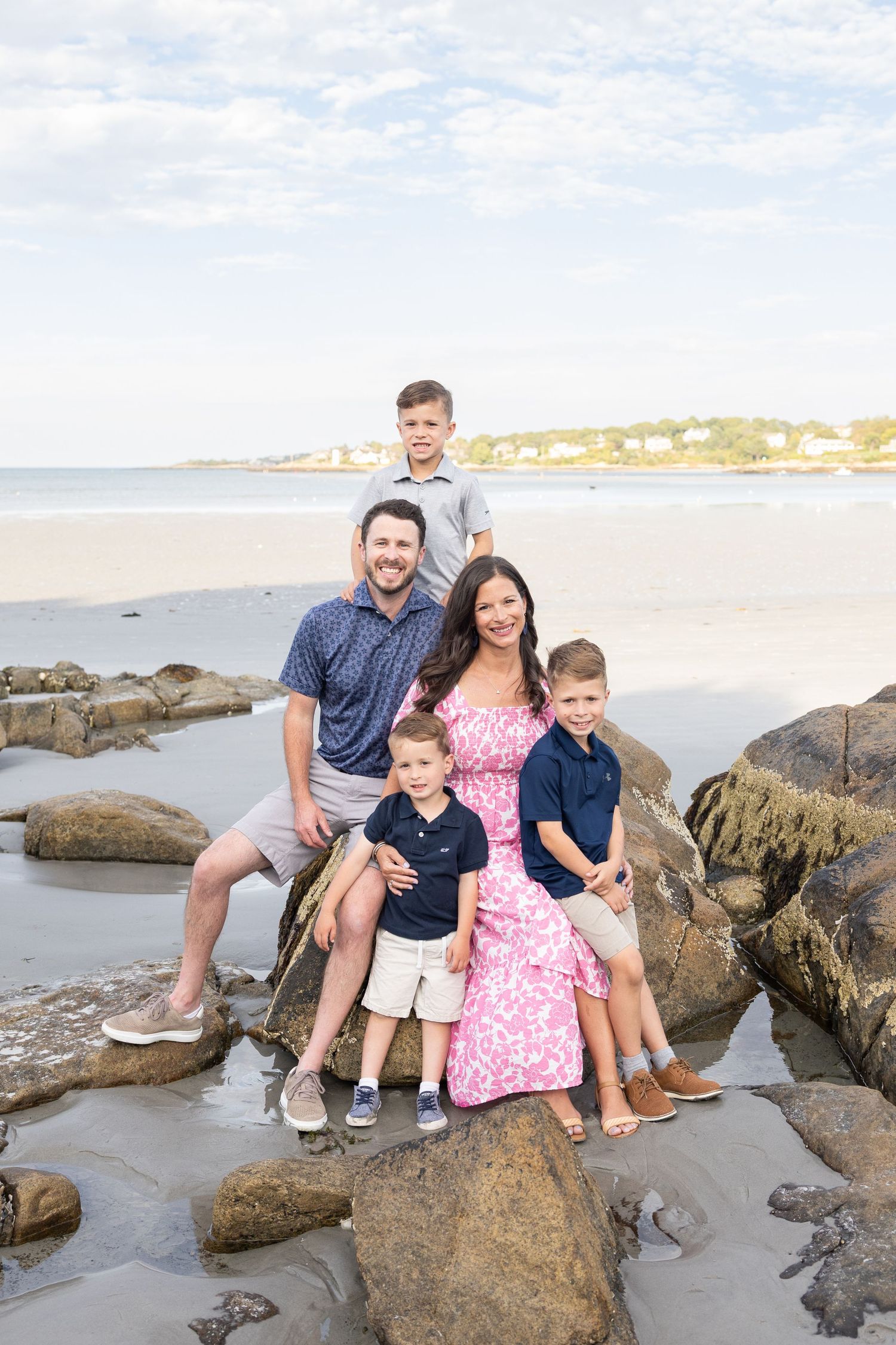 Happy family of five posing on coastal rocks with sandy beach and ocean in background.