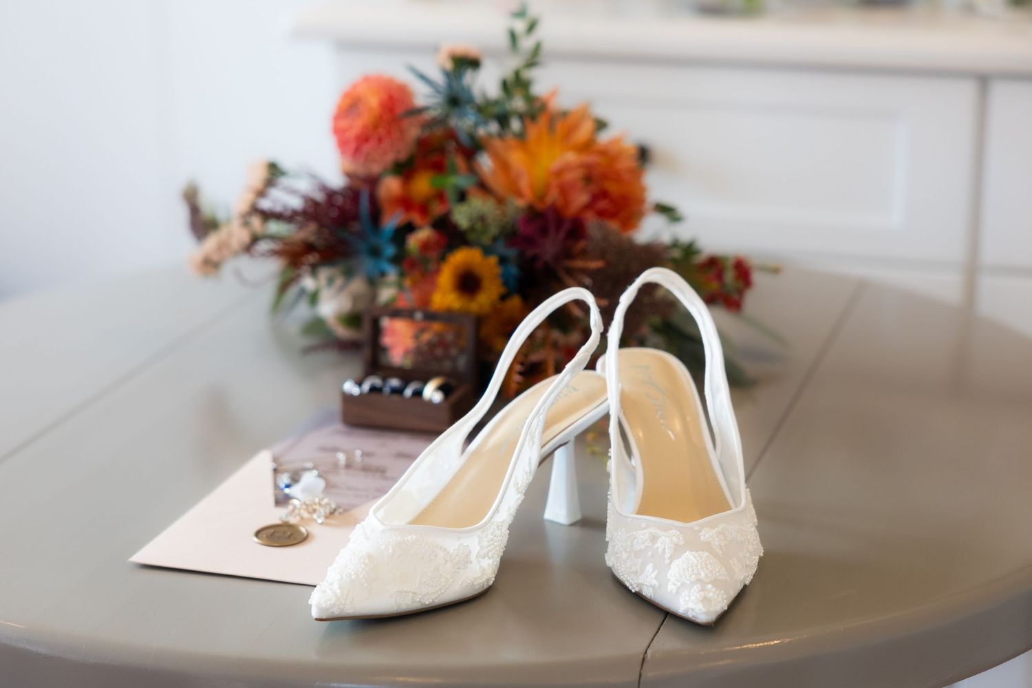 White lace slingback bridal heels displayed on a table with a colorful fall floral arrangement in the background.