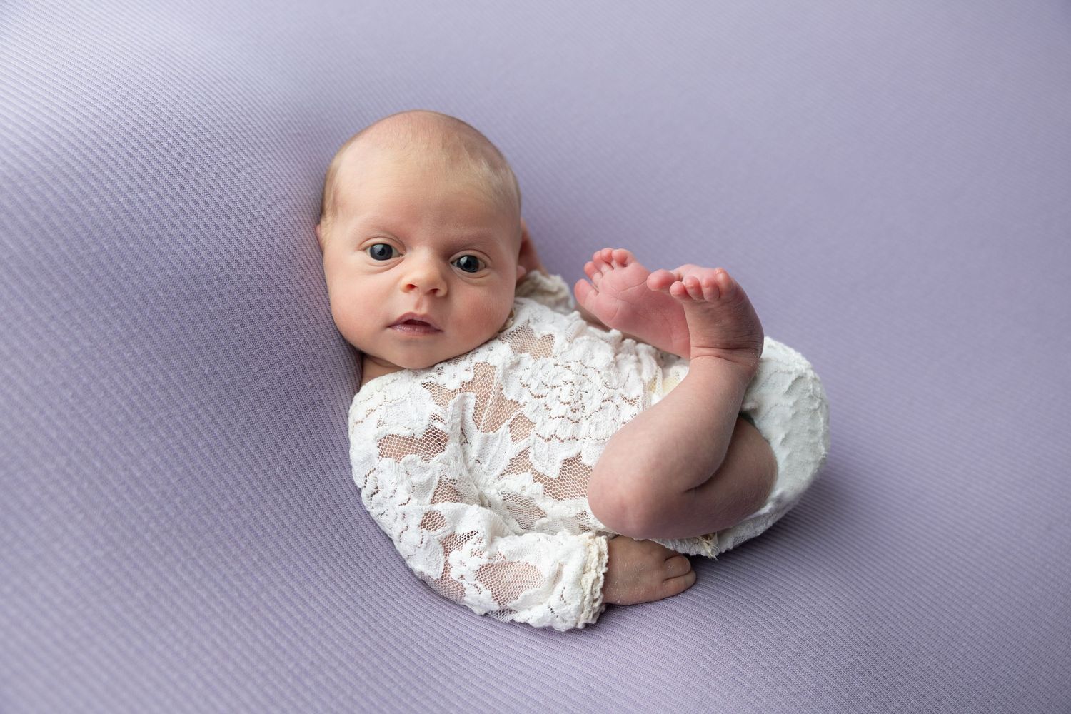 Newborn baby in white lace romper lying on lavender backdrop during professional newborn photography session.
