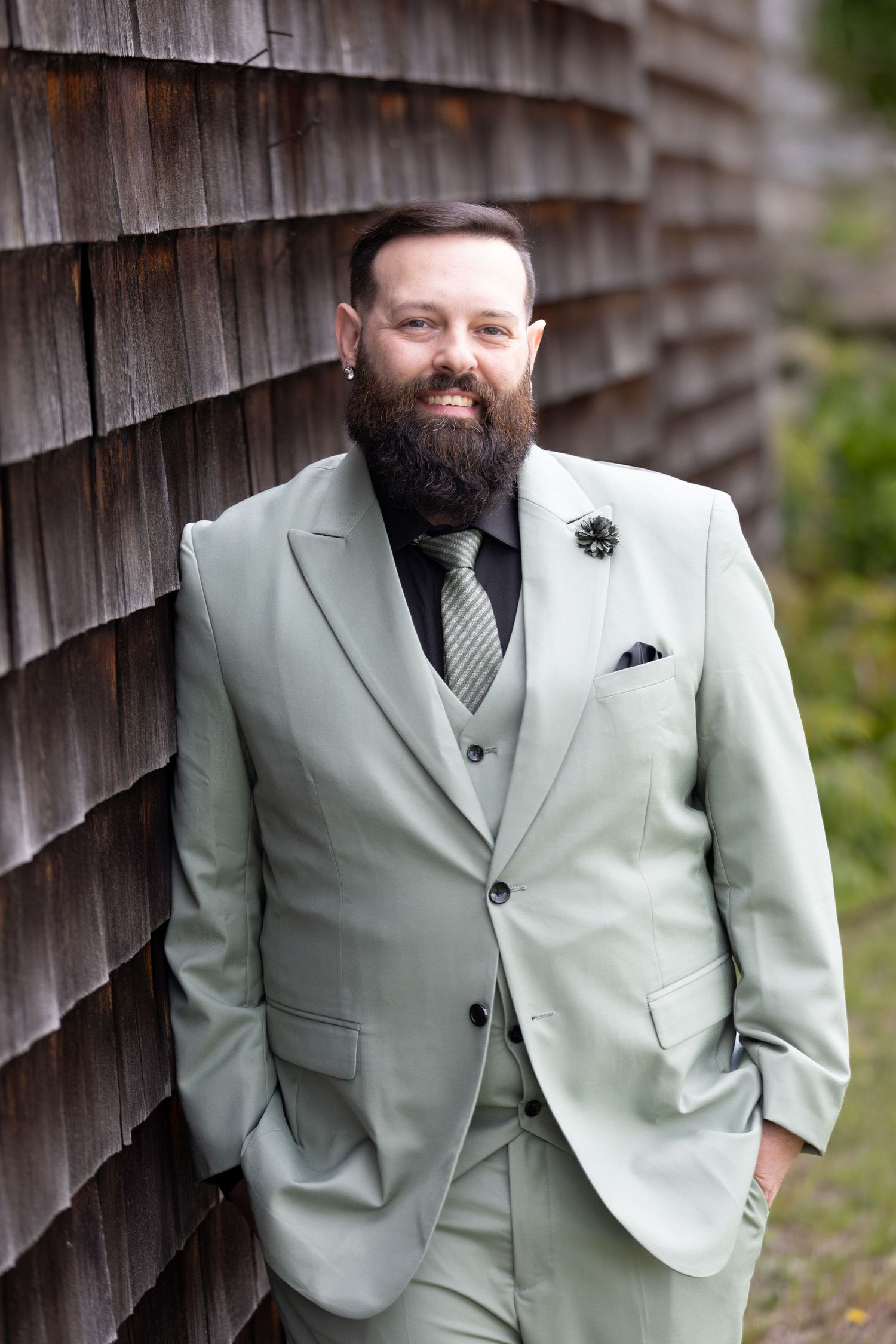 Bearded man in sage green three-piece suit with striped tie leaning against rustic wood shingle wall.