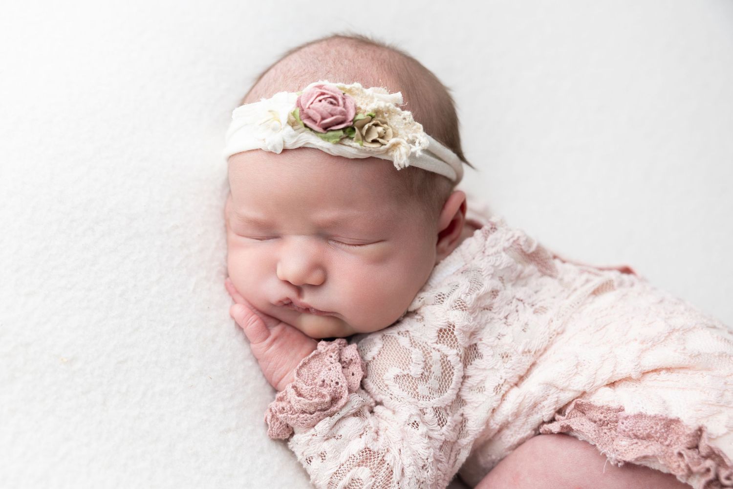 Sleeping newborn baby girl in pink lace romper and floral headband on white blanket.
