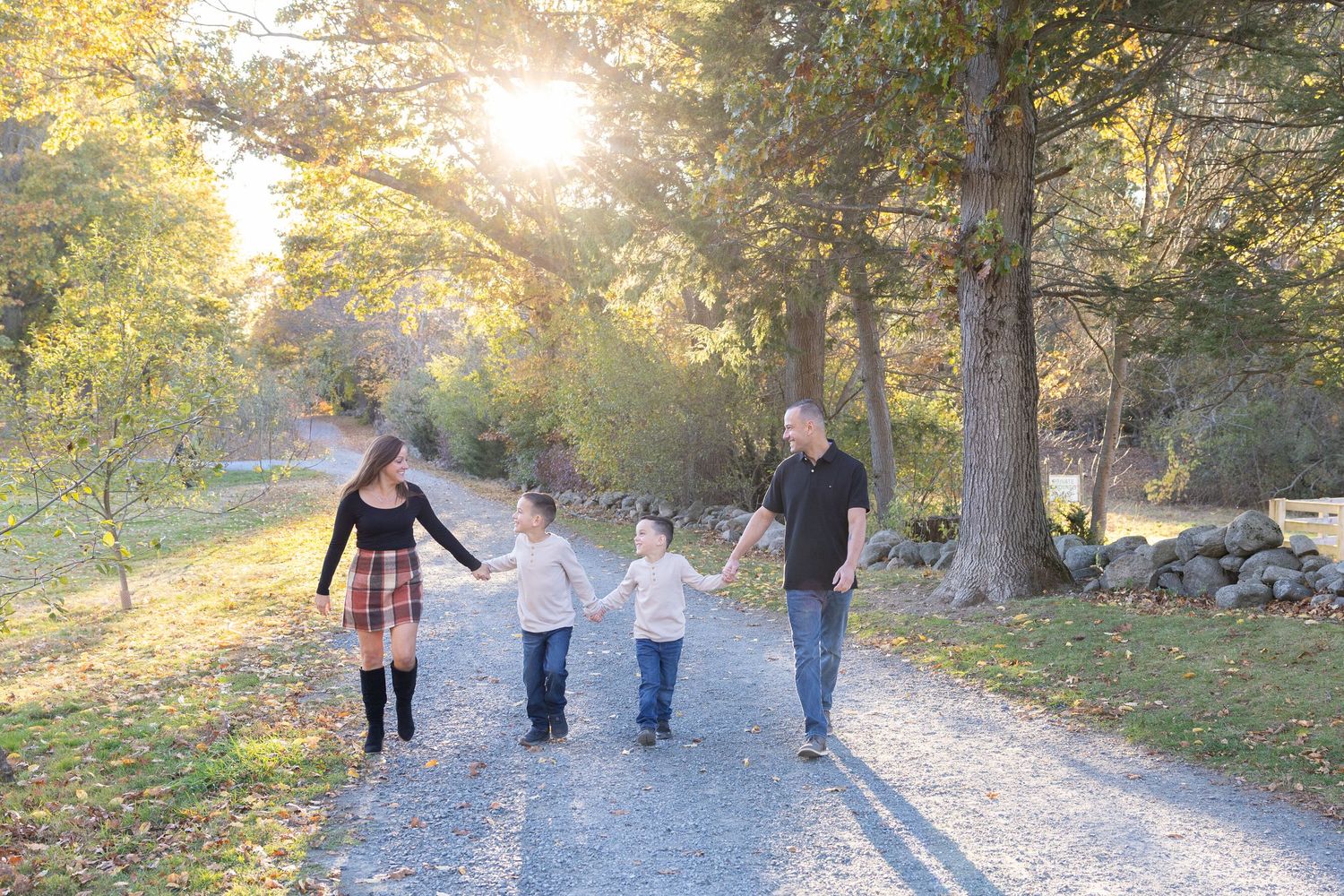 Family of four holding hands walking down sunlit autumn path through golden trees.