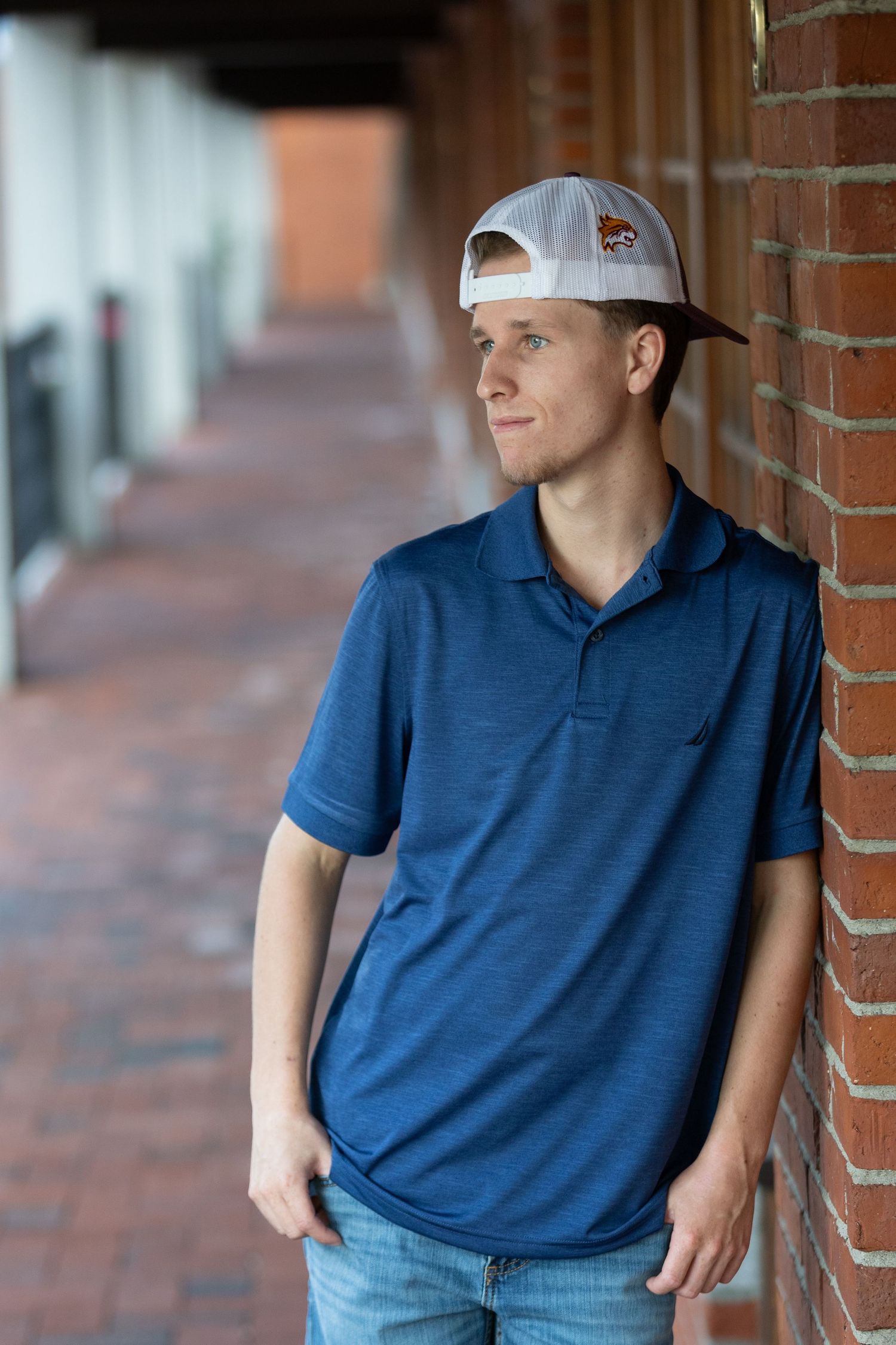 Young man in blue polo shirt and backwards snapback cap leaning against brick wall in outdoor corridor.