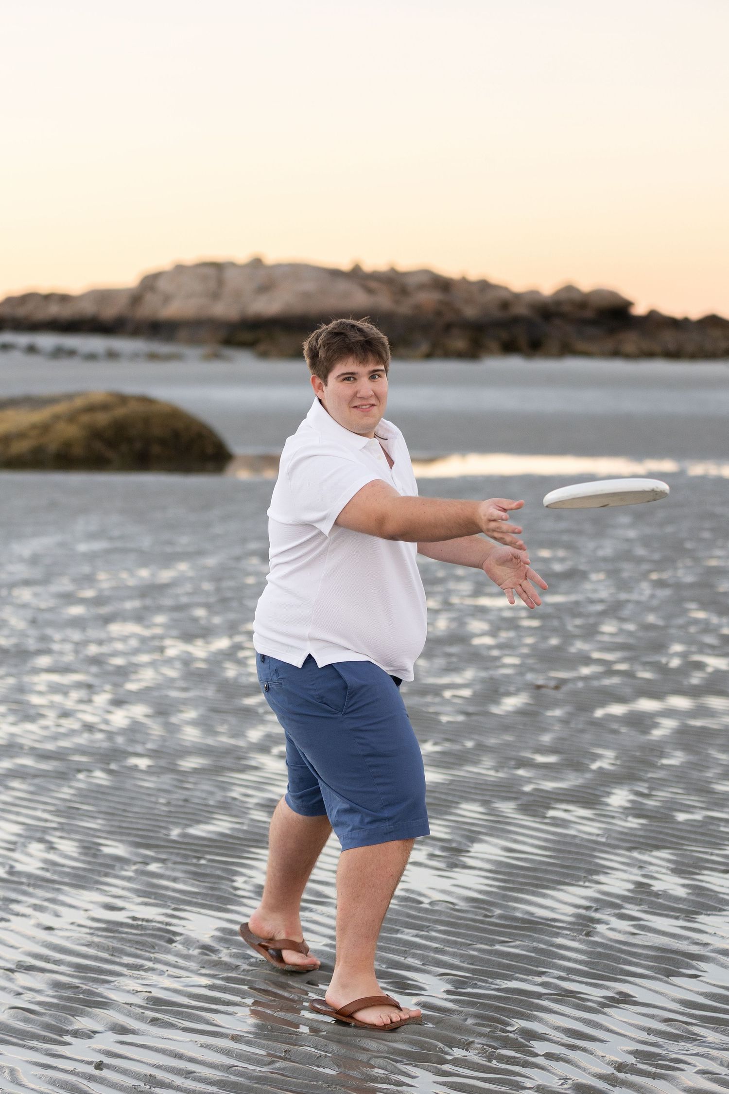 Teen boy in white shirt and blue shorts throwing a white frisbee on a sandy beach at sunset.