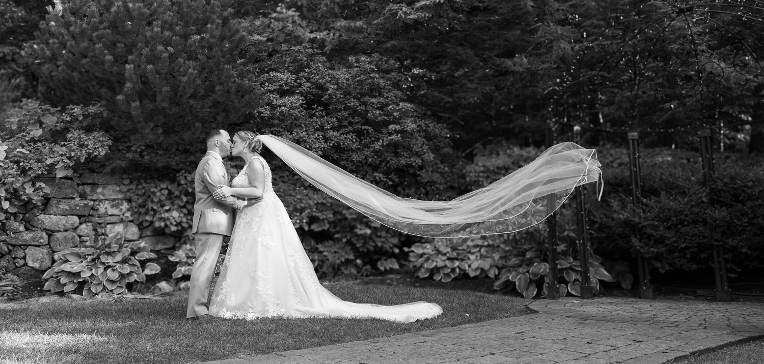 Black and white wedding photo of bride and groom kissing outdoors, with veil flowing dramatically in the wind.