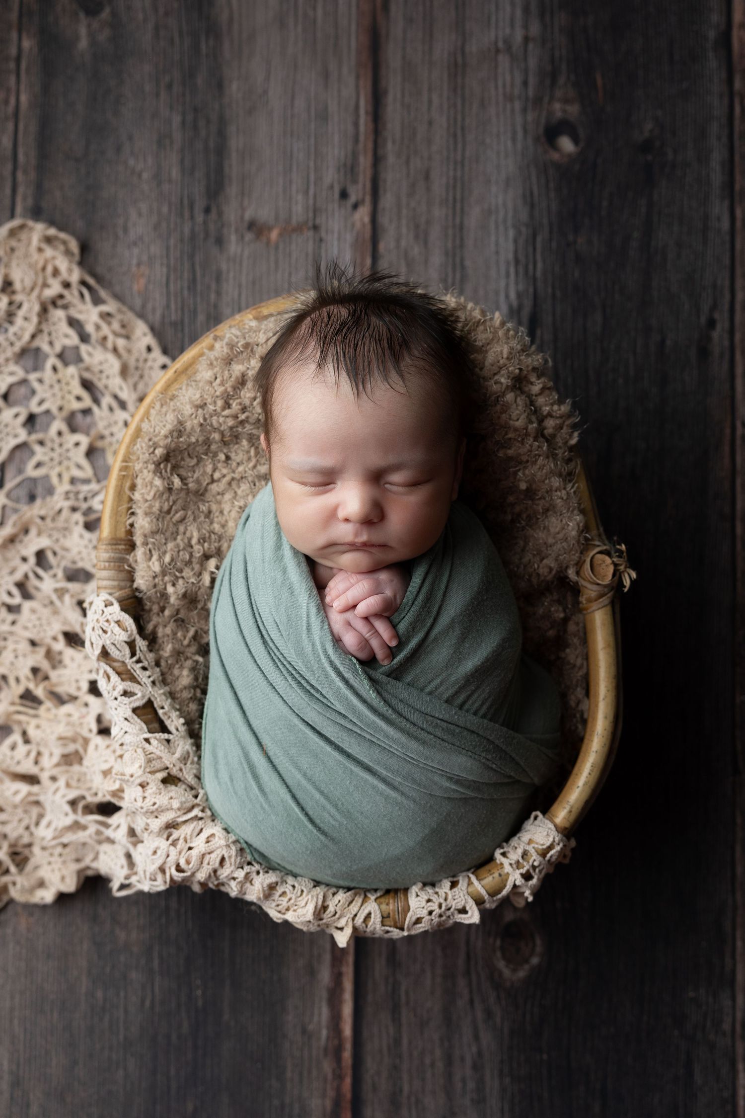 Newborn baby swaddled in teal wrap sleeping peacefully in a round wooden bowl with lace on dark wood.