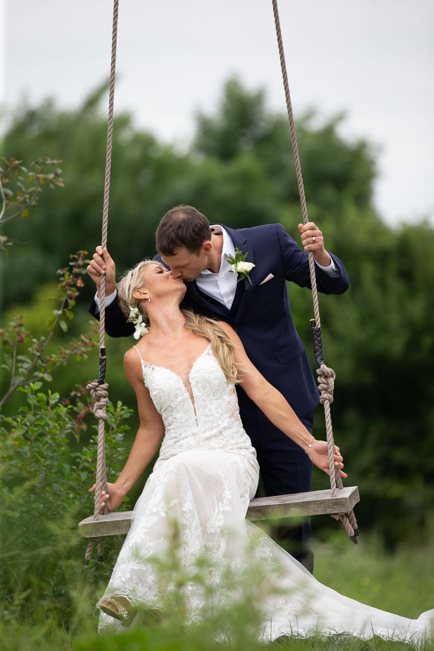 Bride sitting on a wooden rope swing while groom in navy suit leans in for a kiss outdoors.