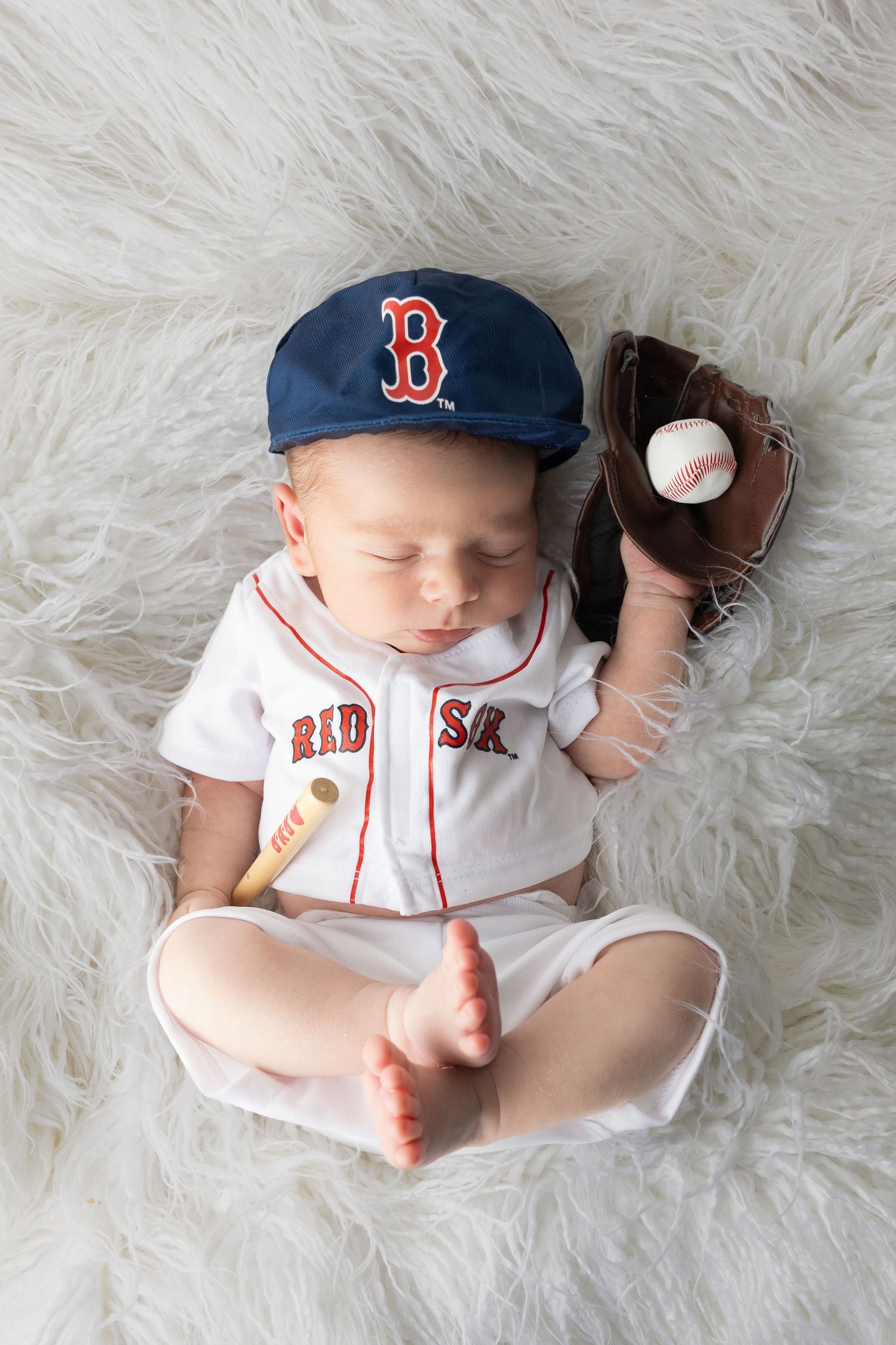 Sleeping newborn baby dressed in Boston Red Sox baseball uniform with cap, mitt, ball, and tiny bat on white fur.