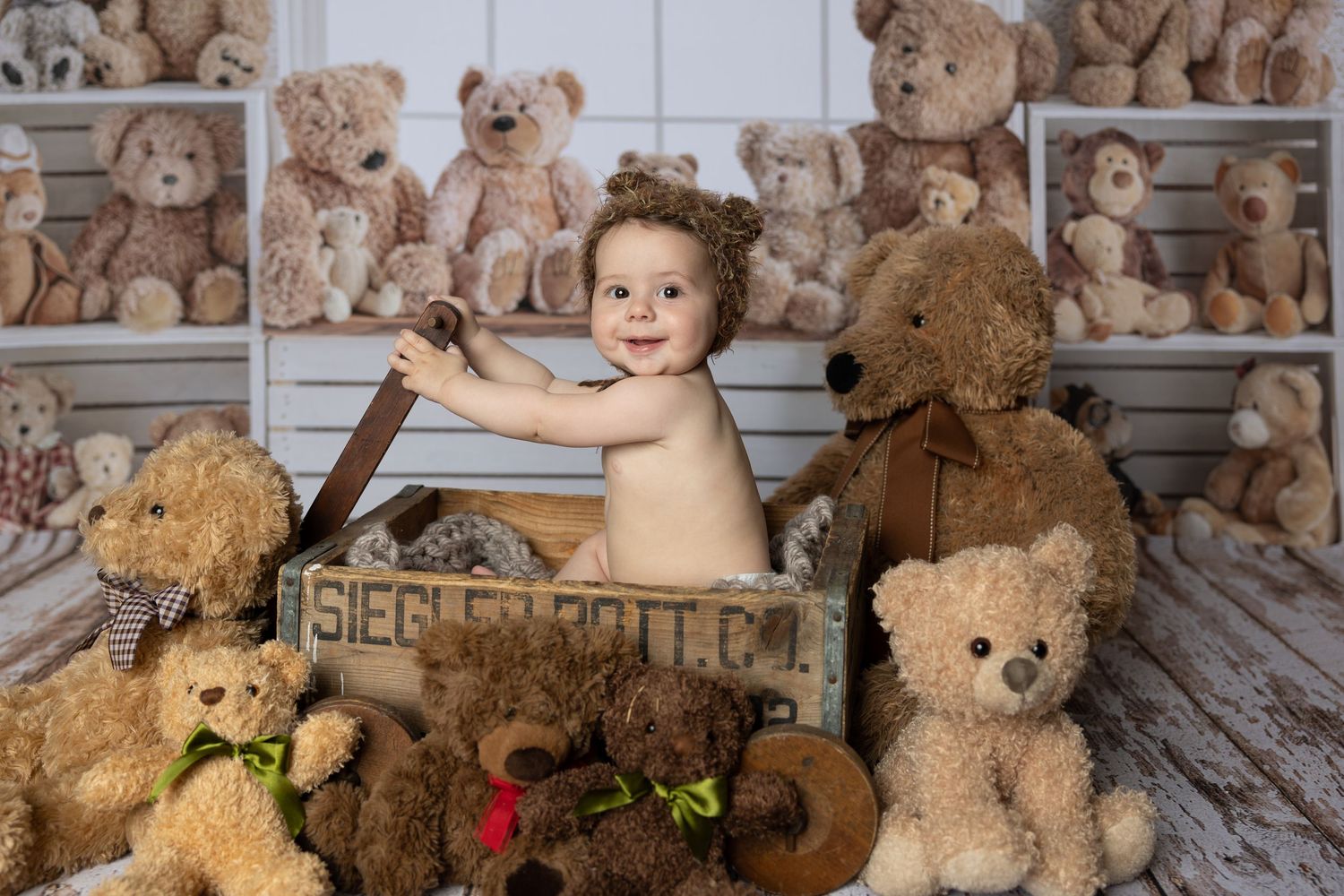 Smiling baby sitting in a wooden crate surrounded by numerous soft teddy bears on white shelving backdrop.
