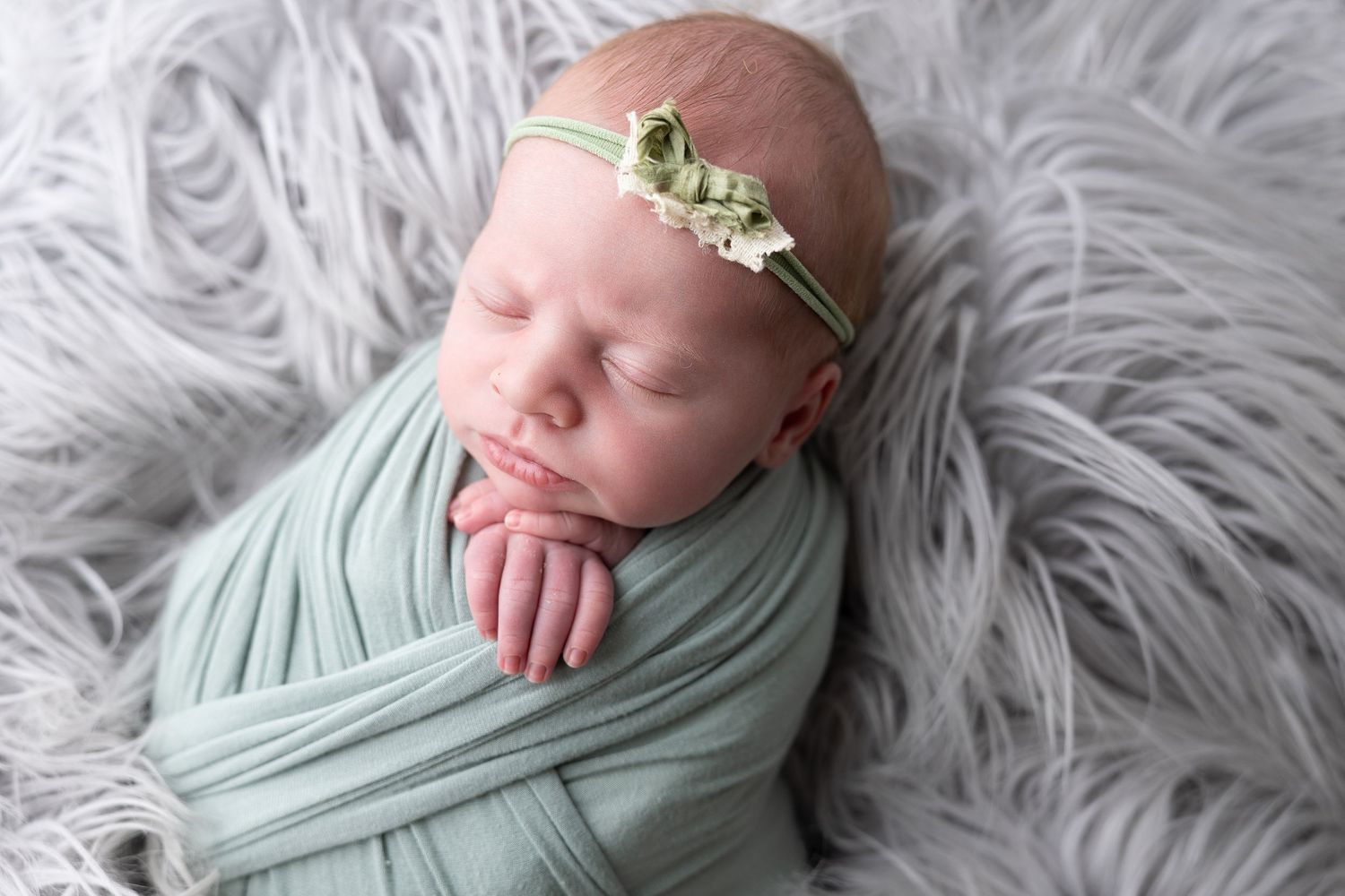 Newborn baby swaddled in mint green wrap with floral headband, sleeping peacefully on fluffy gray fur blanket.