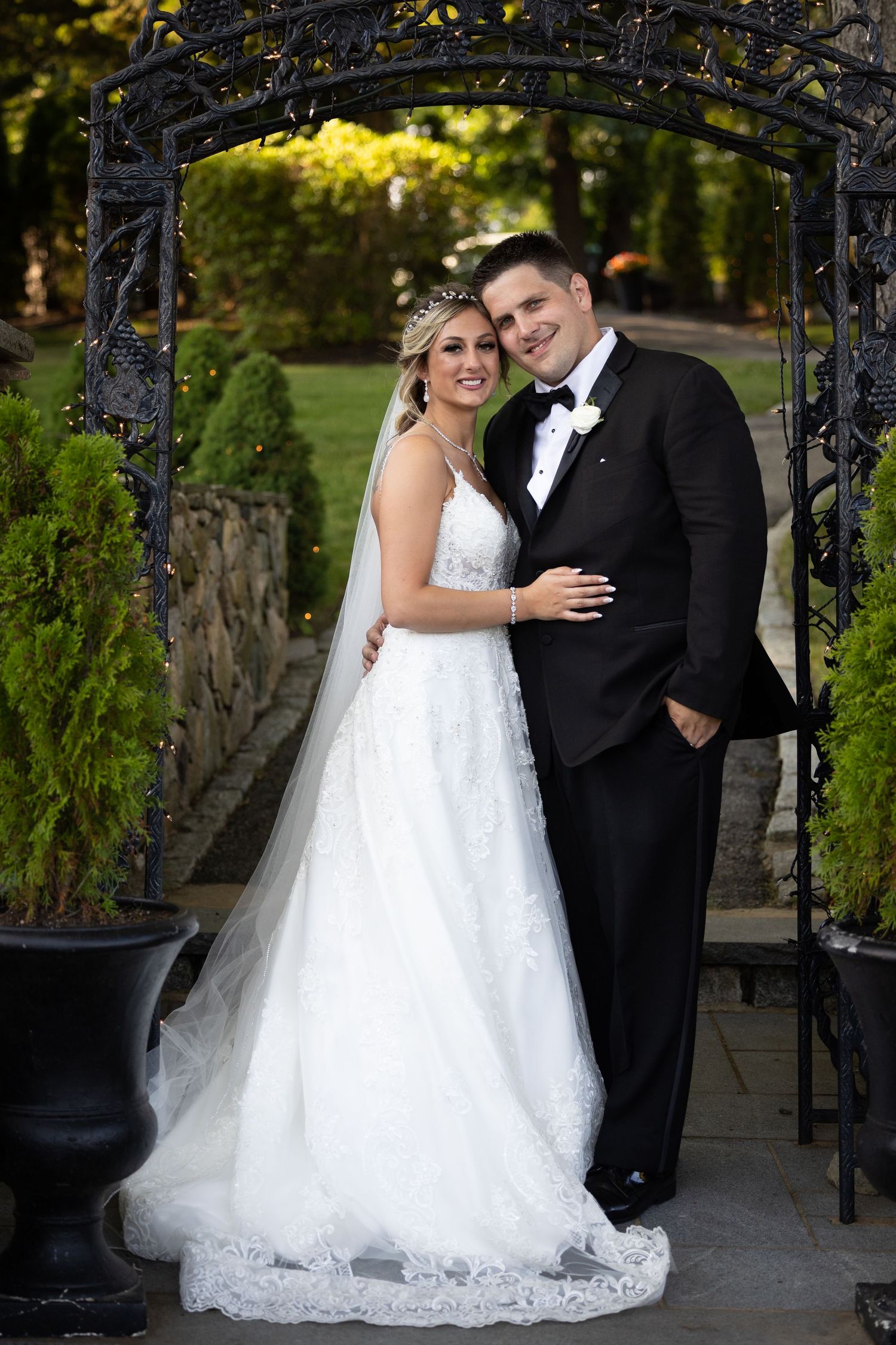 Bride in white lace gown and groom in black tuxedo pose under ornate iron arch in garden setting.