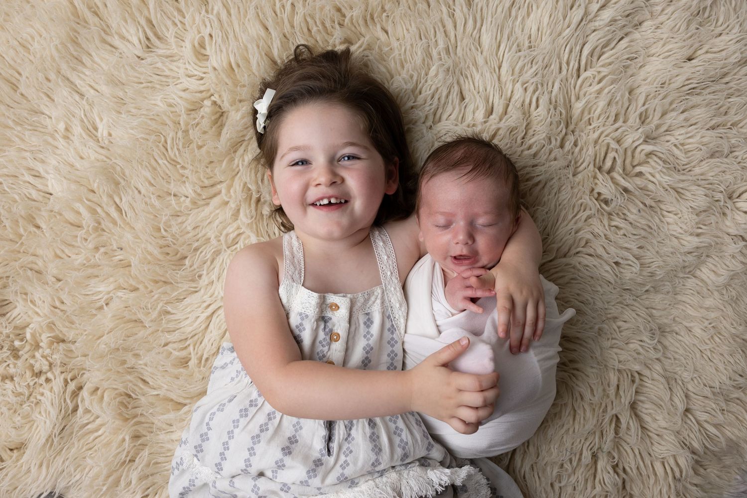 Young girl smiling and hugging a sleeping newborn baby sibling on a fluffy cream rug.