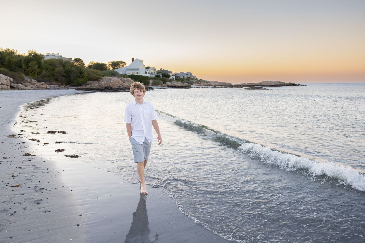 A boy in white shirt and shorts walks barefoot along a sandy beach at sunset with calm ocean waves.