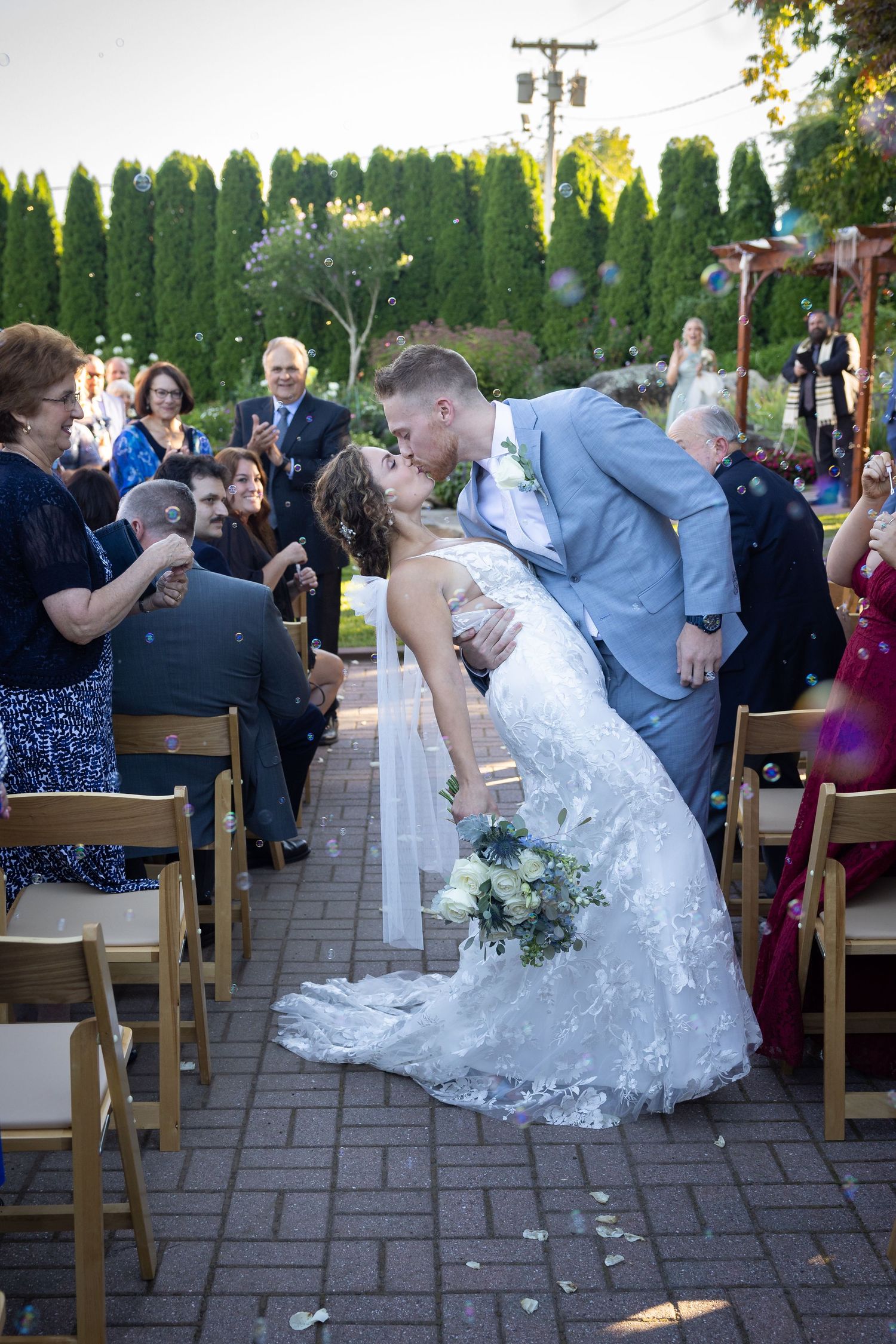 Bride and groom share a romantic kiss at outdoor wedding ceremony while guests celebrate with bubbles.