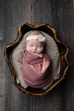 Serene newborn baby swaddled in dusty rose wrap resting in a rustic wooden bowl.