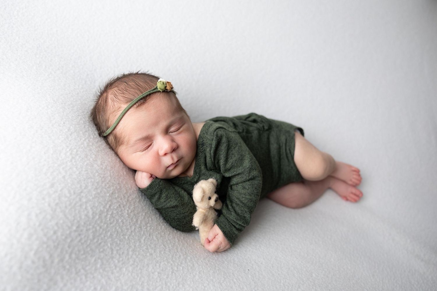 Sleeping newborn baby in green romper holding tiny teddy bear on white blanket with floral headband.