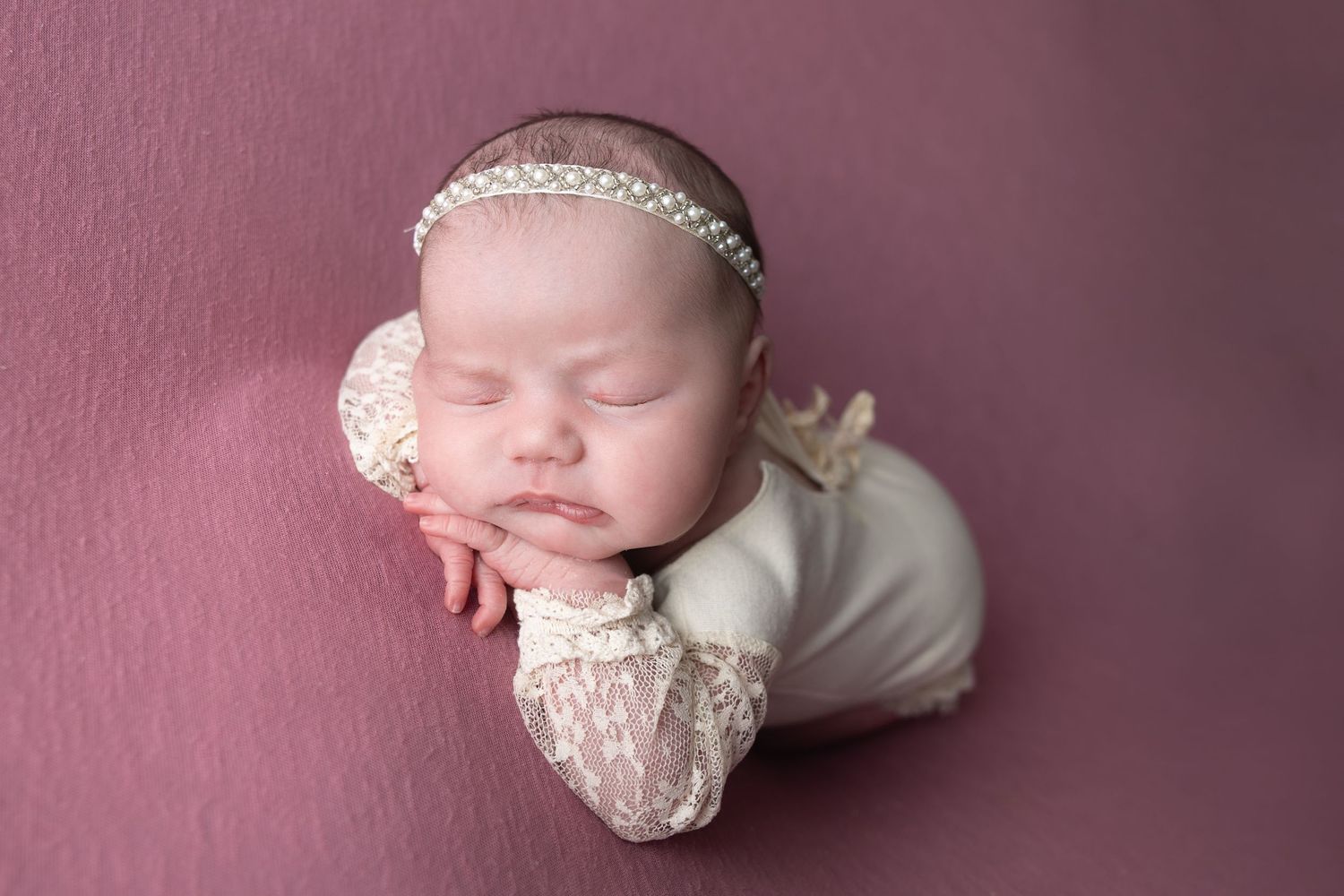 Newborn baby girl sleeping peacefully in lace romper and pearl headband on mauve pink backdrop.