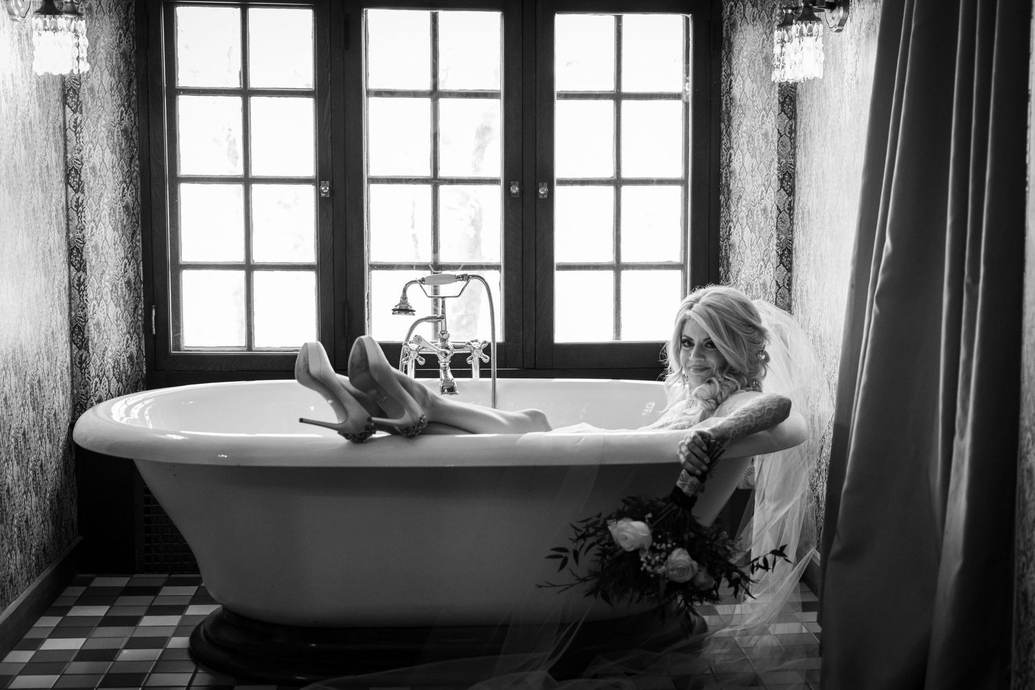 Bride in wedding dress and veil sitting in a vintage clawfoot bathtub, black and white photo with French windows.