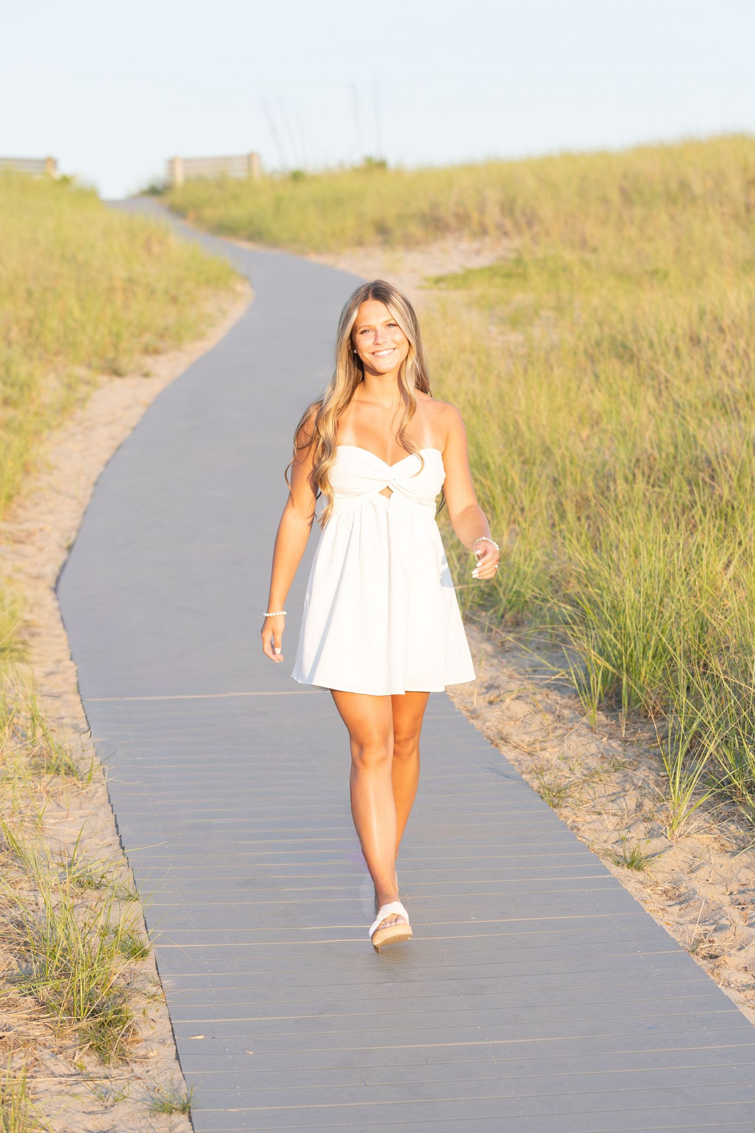 Young woman in white sundress walking on wooden boardwalk path through beach dunes on sunny day.