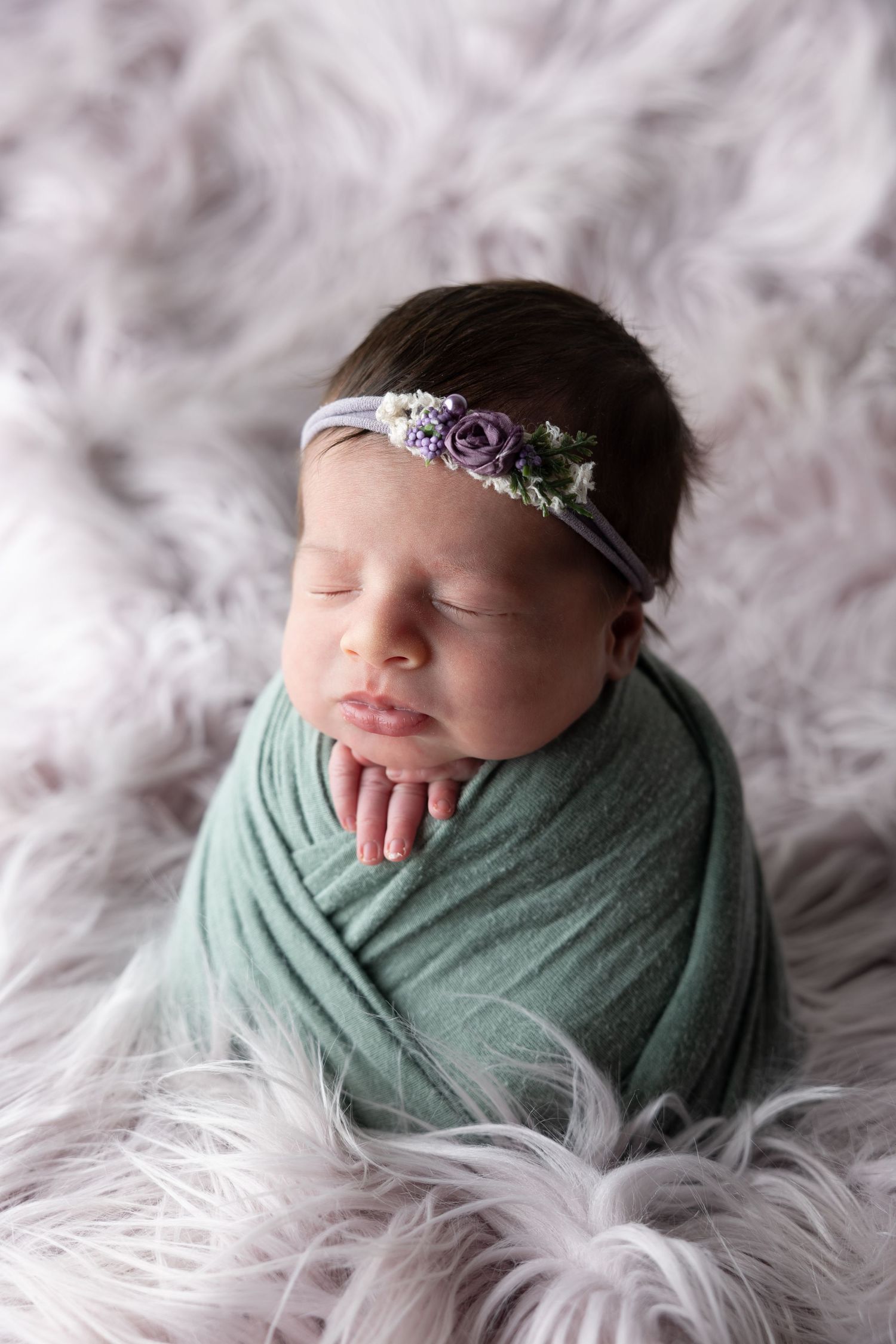 Sleeping newborn baby wrapped in green swaddle with purple floral headband on fluffy lavender fur backdrop.