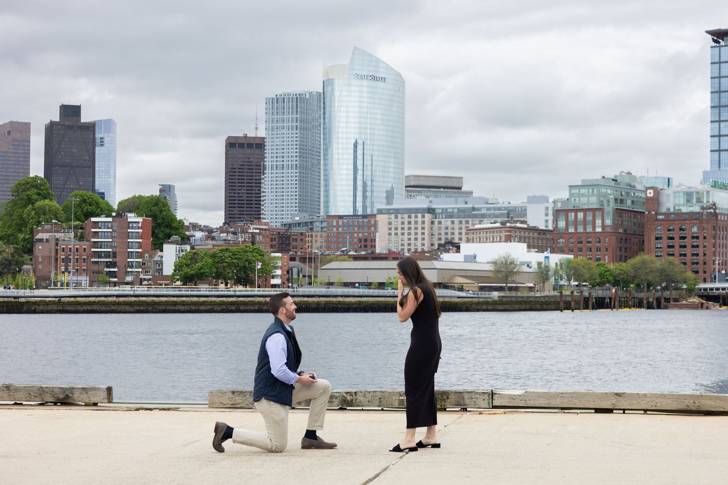 Man proposes on one knee to woman by Boston Harbor waterfront with city skyline backdrop.