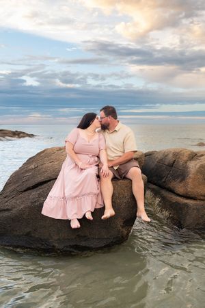Couple sitting closely on ocean rocks sharing a tender moment at sunset with calm water below.