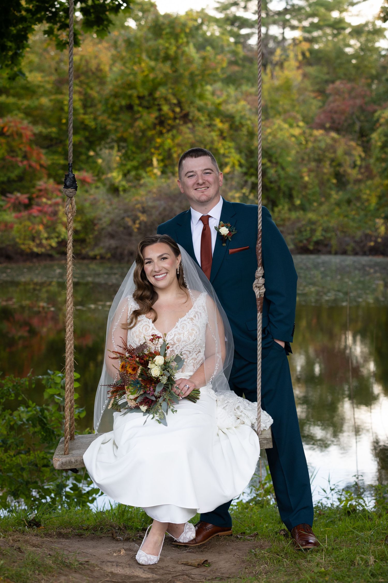 Bride sitting on rope swing holding autumn bouquet with groom standing behind her, posing near a pond with fall foliage.