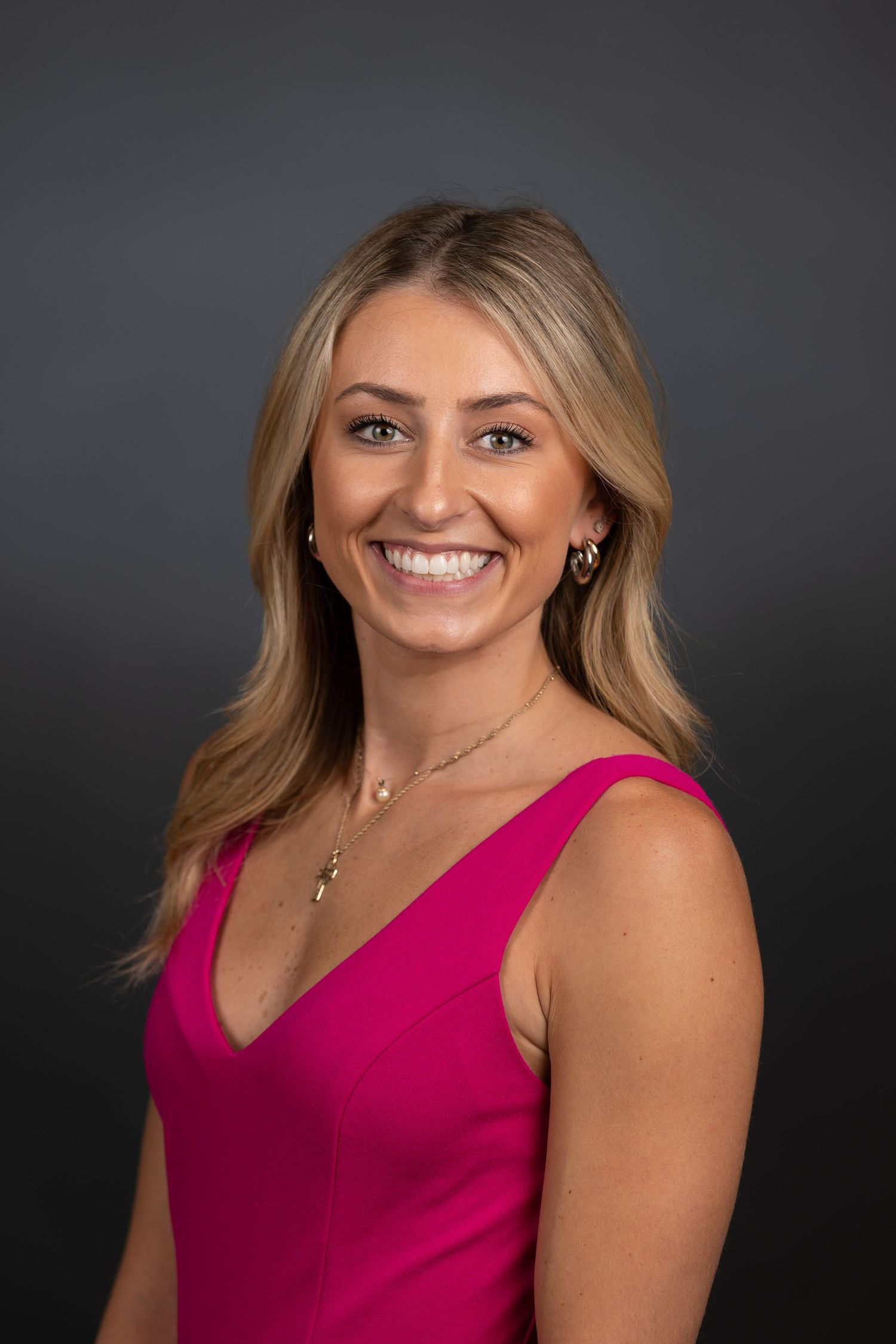 Professional headshot of a smiling blonde woman in a pink dress against a dark gray background.