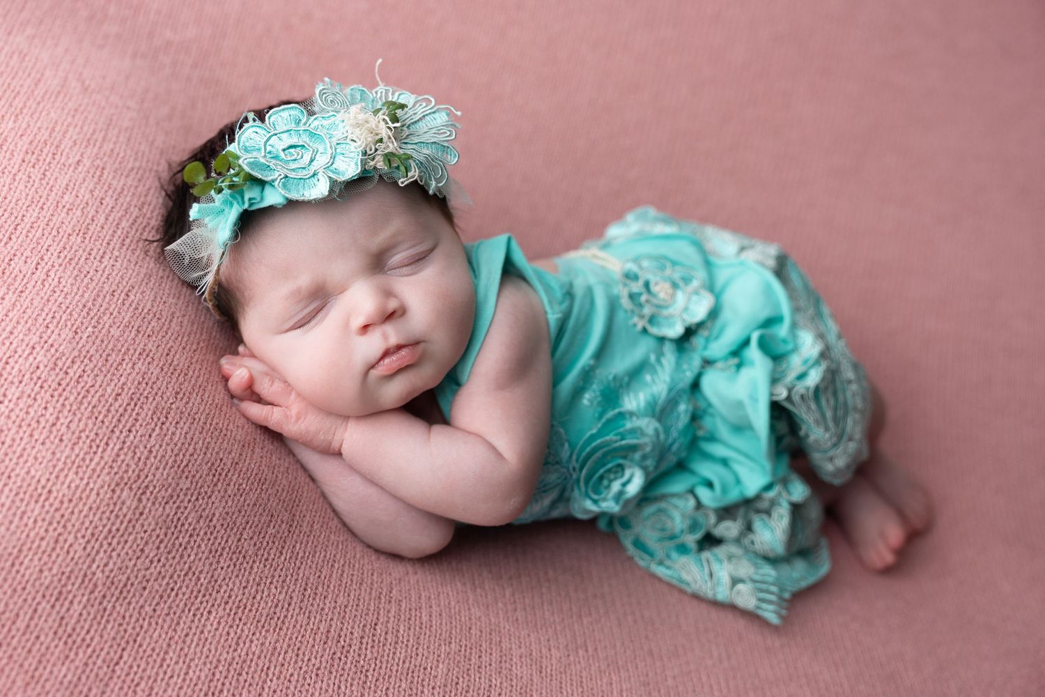 Sleeping newborn baby girl in teal floral outfit and matching flower headband on pink backdrop.