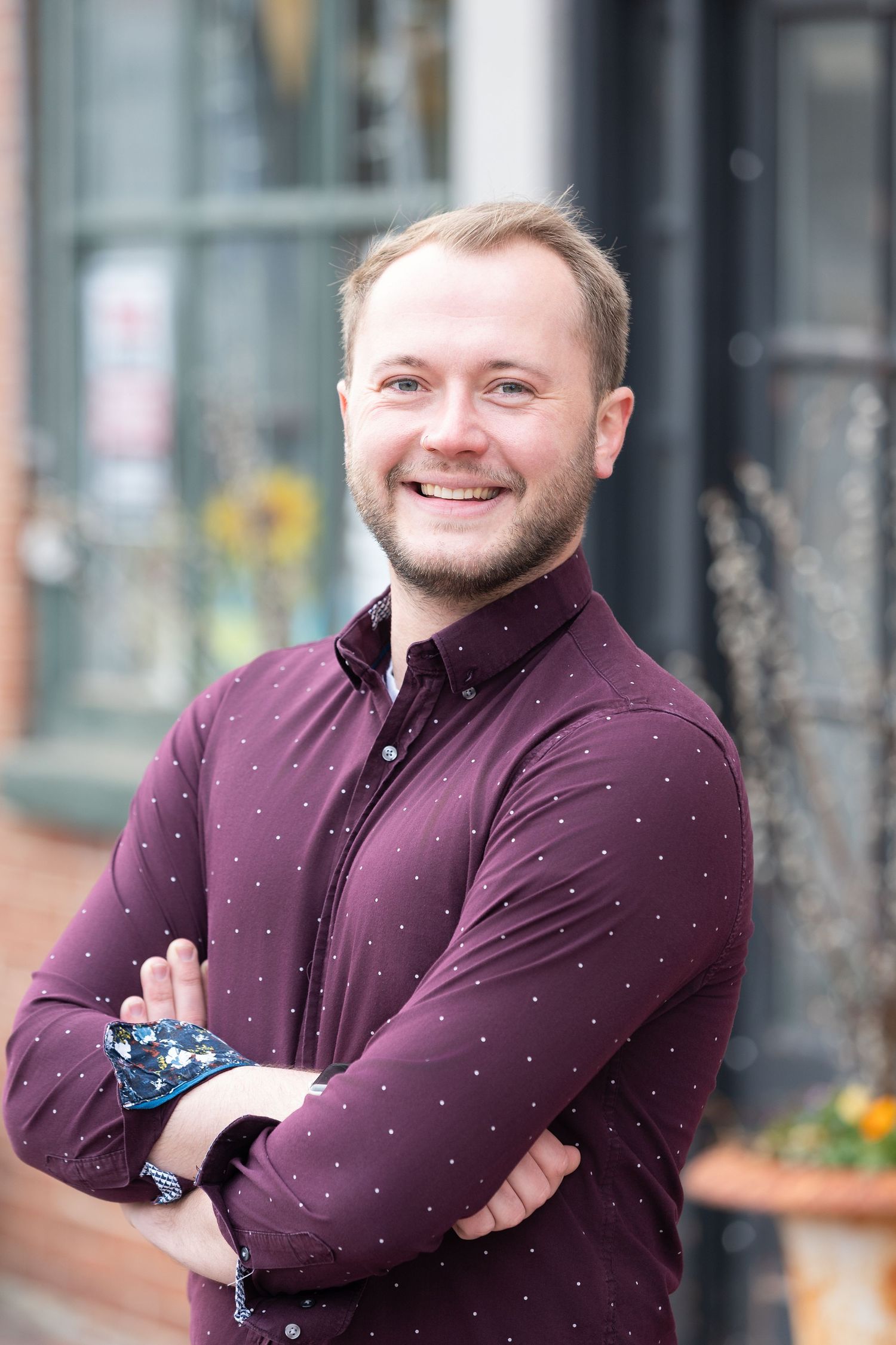 Smiling young man with beard, arms crossed, wearing a maroon polka dot shirt, standing outdoors in an urban setting.