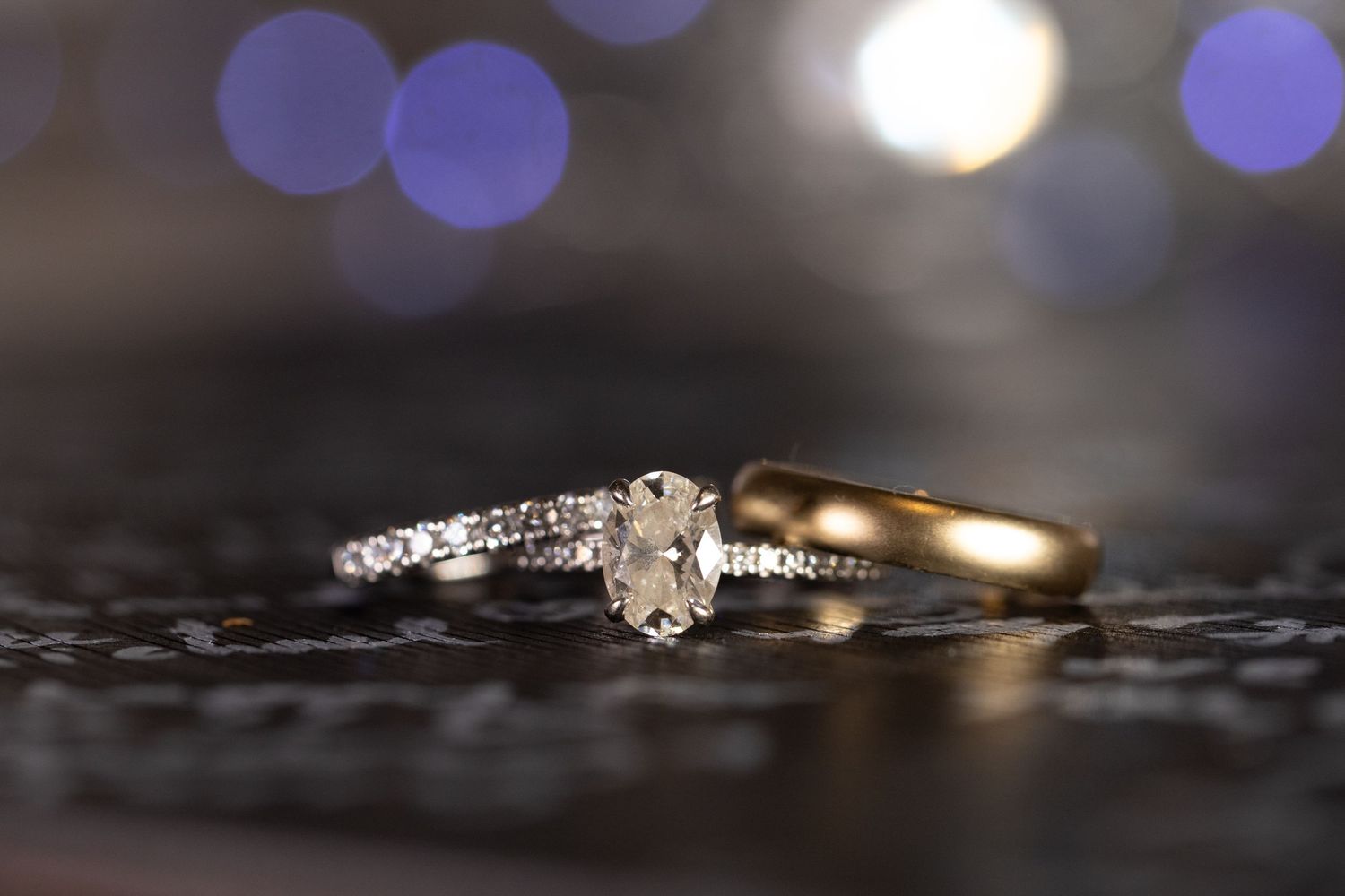 Elegant diamond engagement ring paired with a gold wedding band on dark surface with soft bokeh lights.