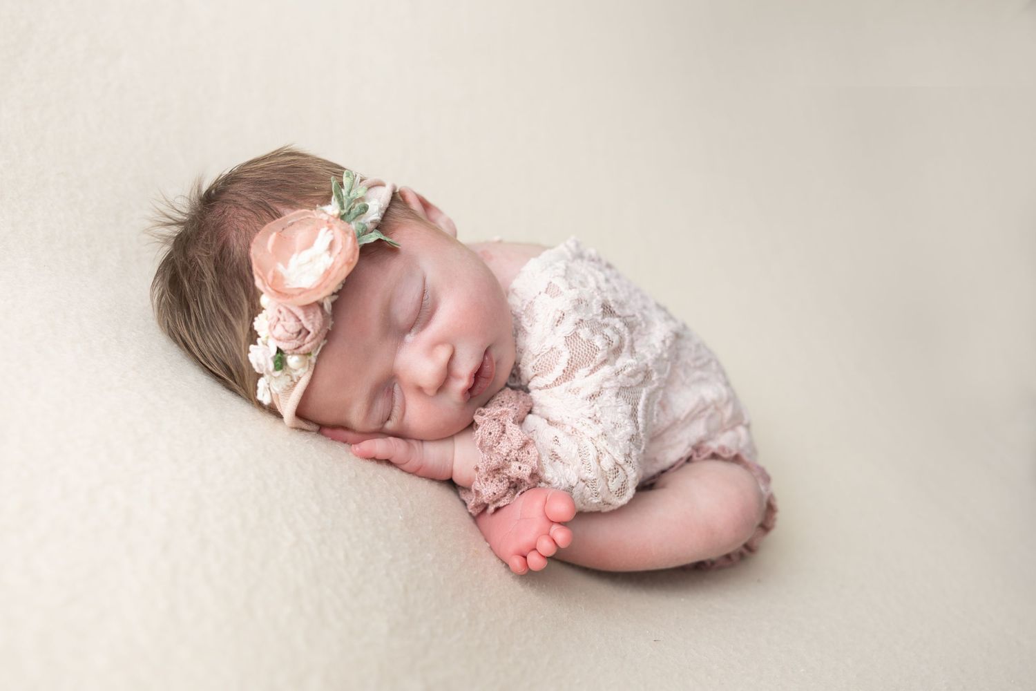 Sleeping newborn baby girl in lace romper and floral headband on cream blanket.
