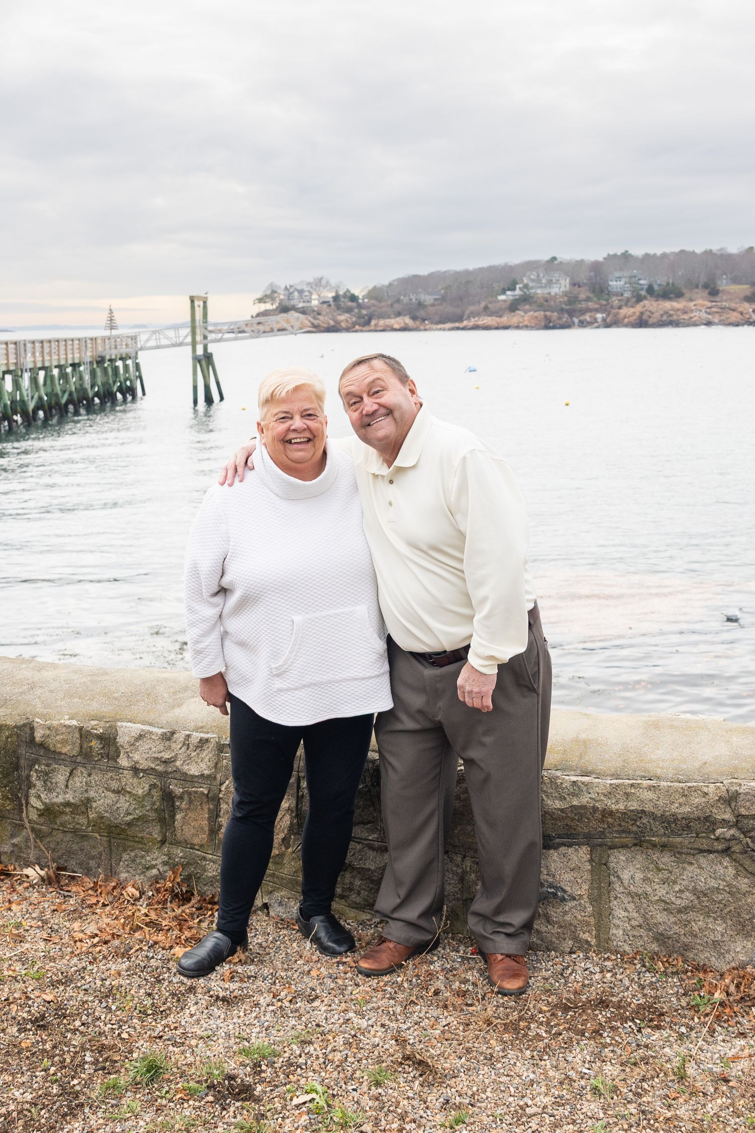 Smiling senior couple embracing by waterfront stone wall on overcast day near marina pier.