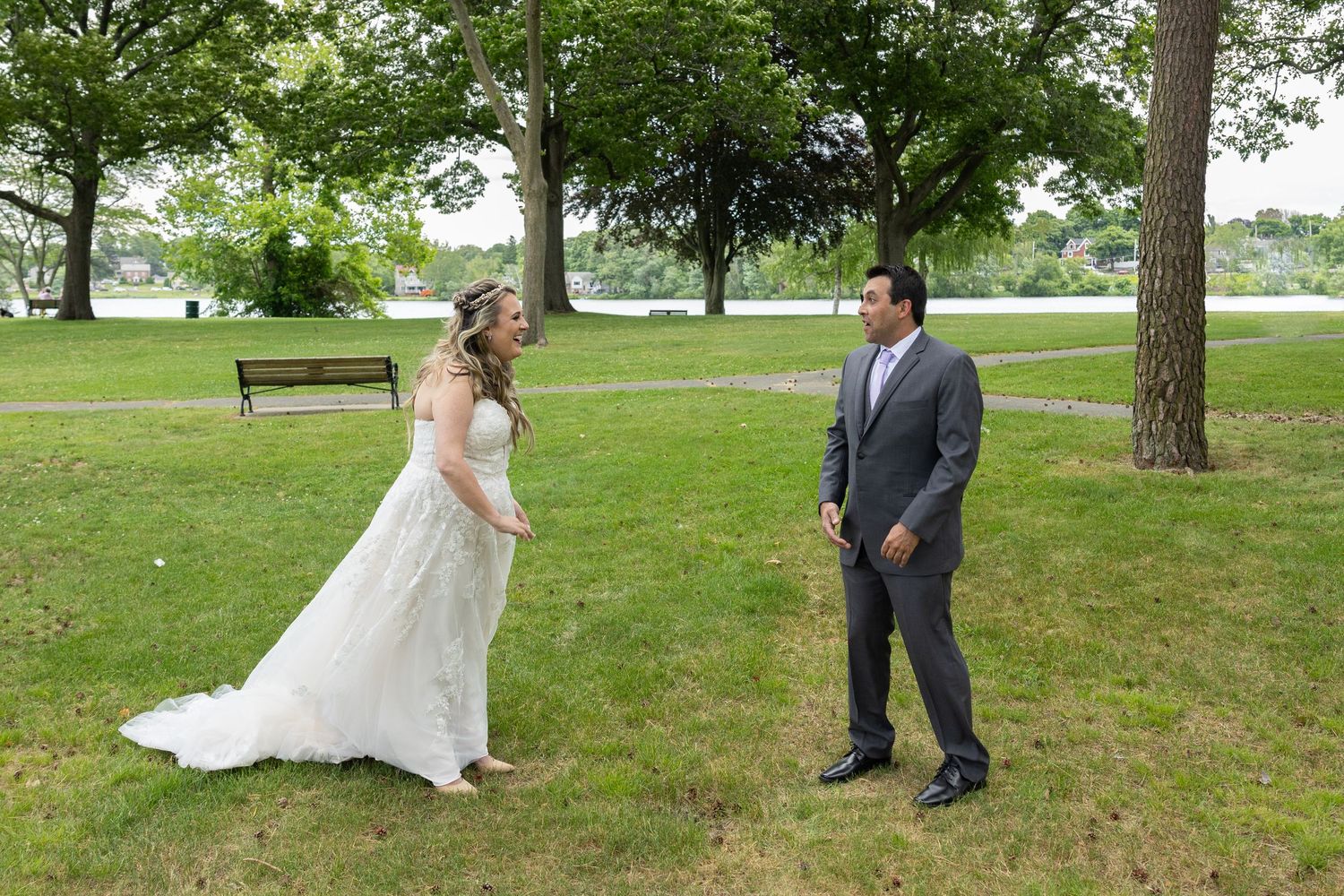 Bride in white gown and groom in gray suit share a joyful first look moment in a lush green park.