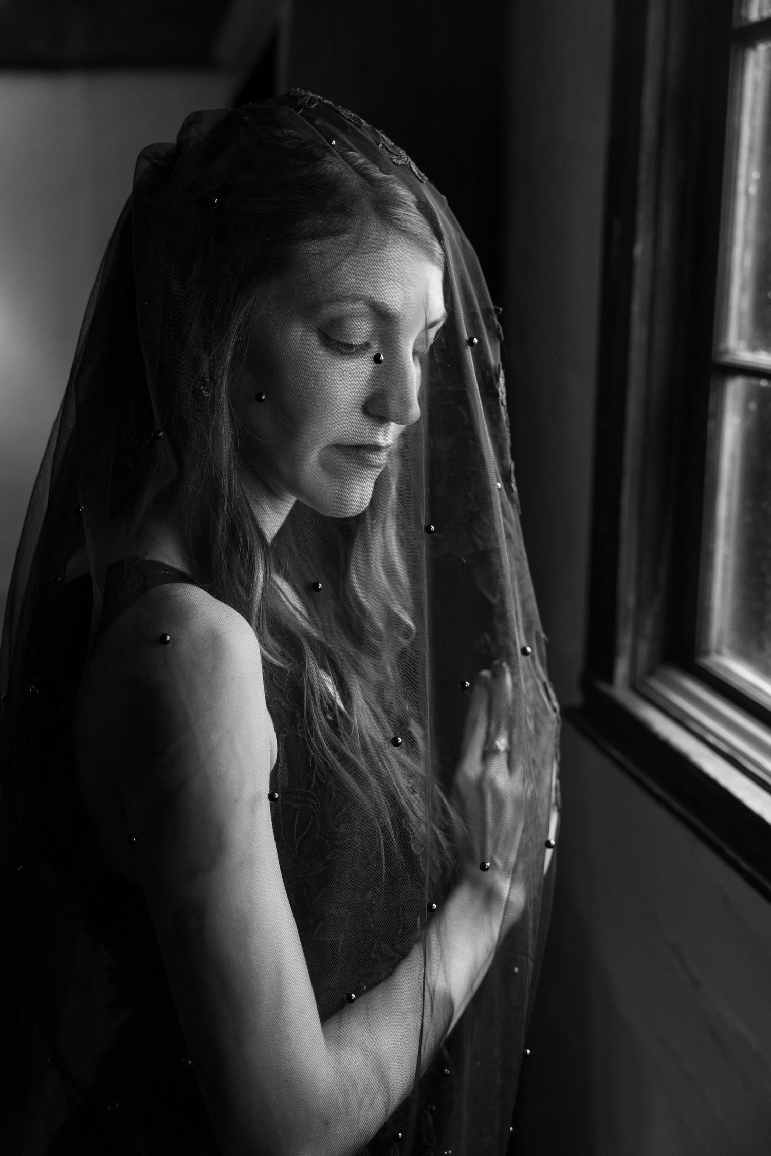Black and white portrait of a bride wearing a pearl-studded veil, gazing down by a window in soft light.