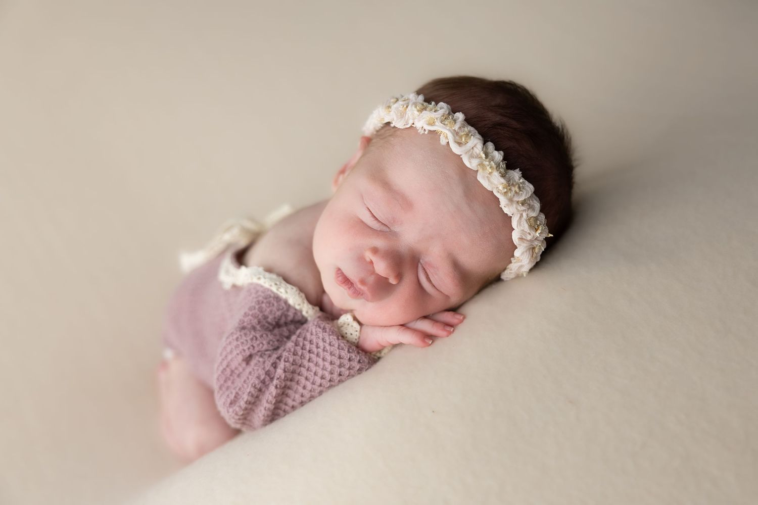 Sleeping newborn baby girl in purple romper and white floral headband on cream background.