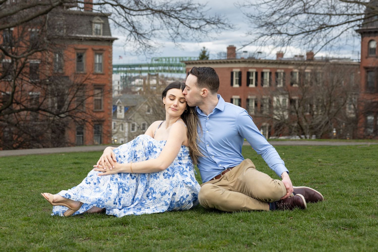 Couple sitting together on green grass in a city park with historic brick buildings in background.
