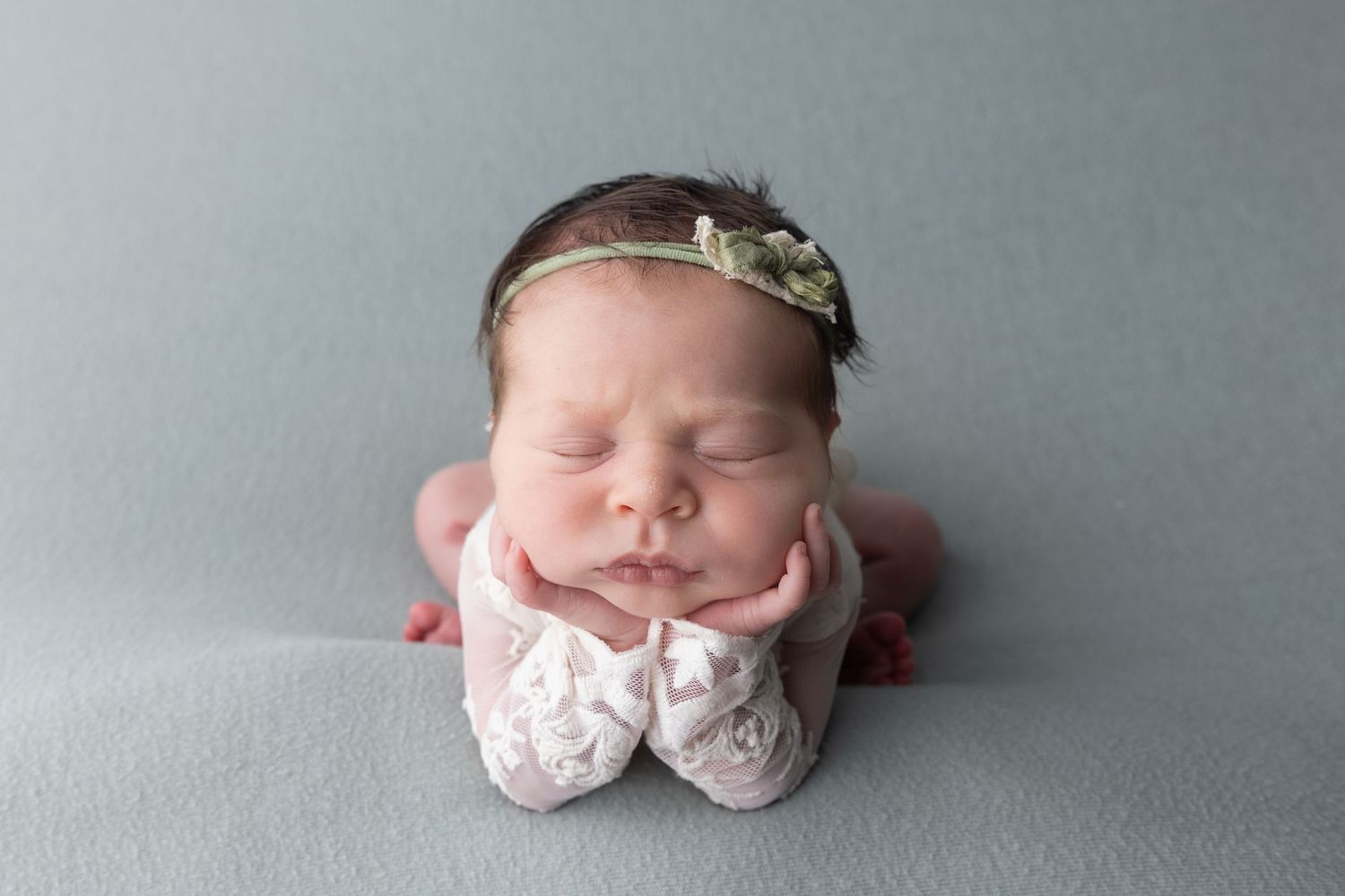 Sleeping newborn baby girl in froggy pose wearing floral headband and white lace booties on blue backdrop.