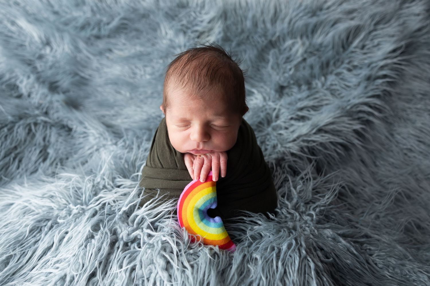 Newborn baby wrapped in dark green swaddle holding a colorful rainbow toy on a soft blue fur blanket.