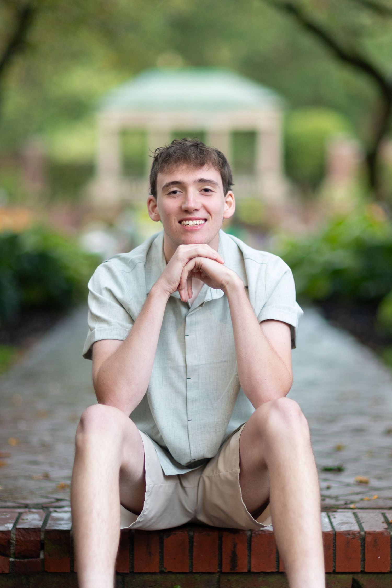 Smiling young man sitting on brick steps in a garden pathway with a gazebo in the background.