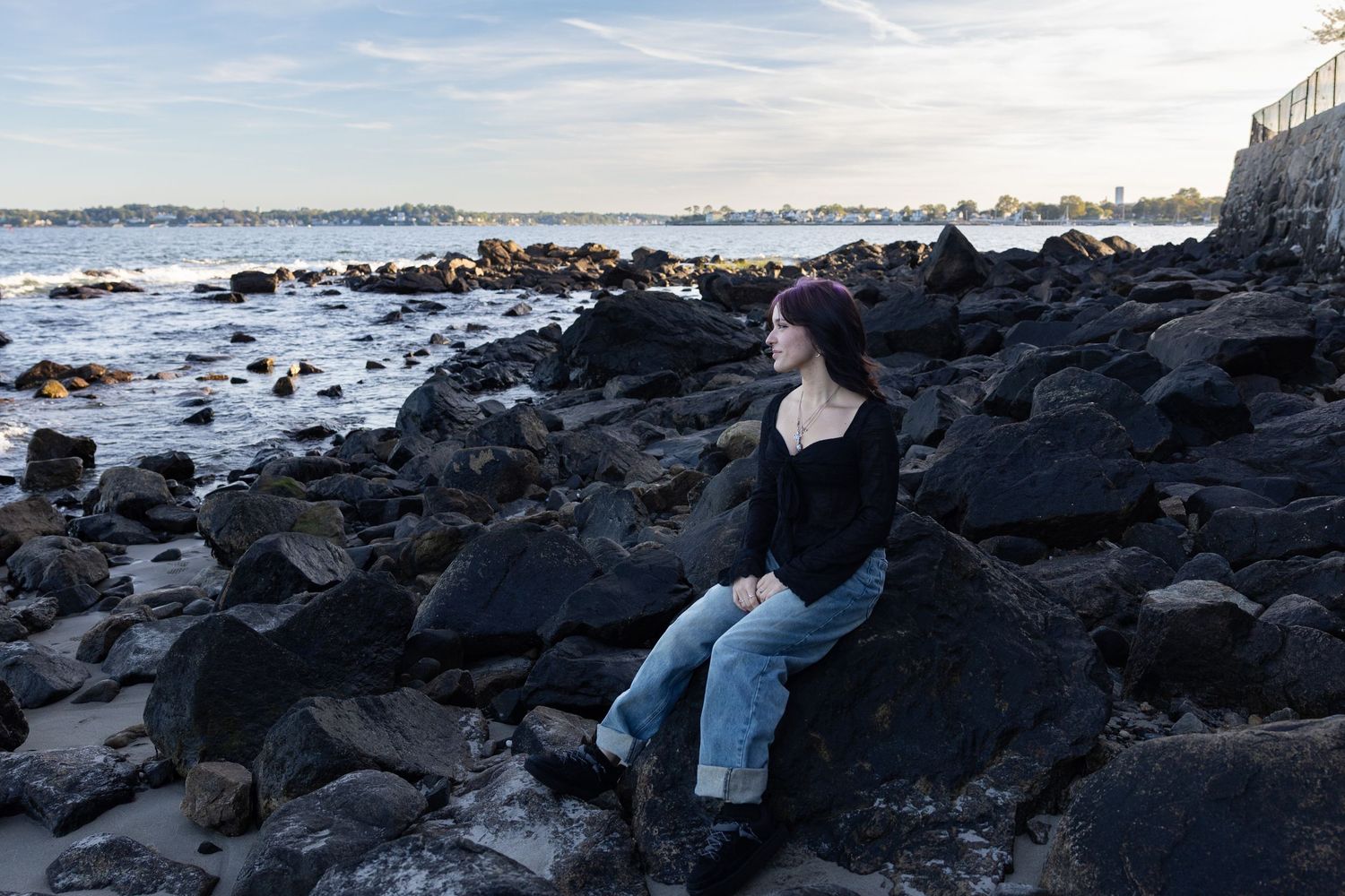 Young woman in black top and jeans sitting on rocky shoreline, gazing at ocean waves under a cloudy sky.