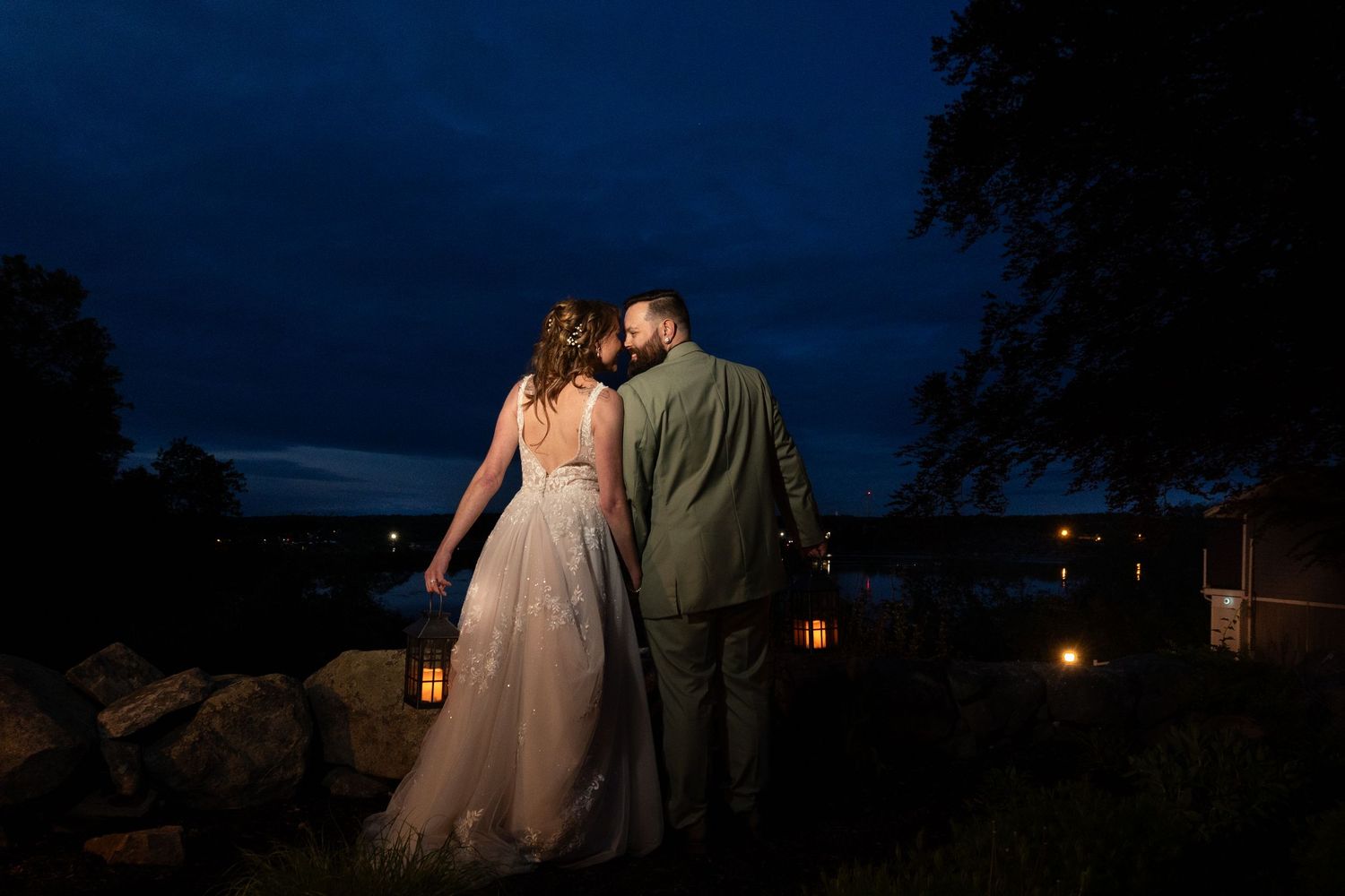 Bride and groom share a romantic moment at dusk, framed by glowing lanterns with a serene waterfront backdrop.