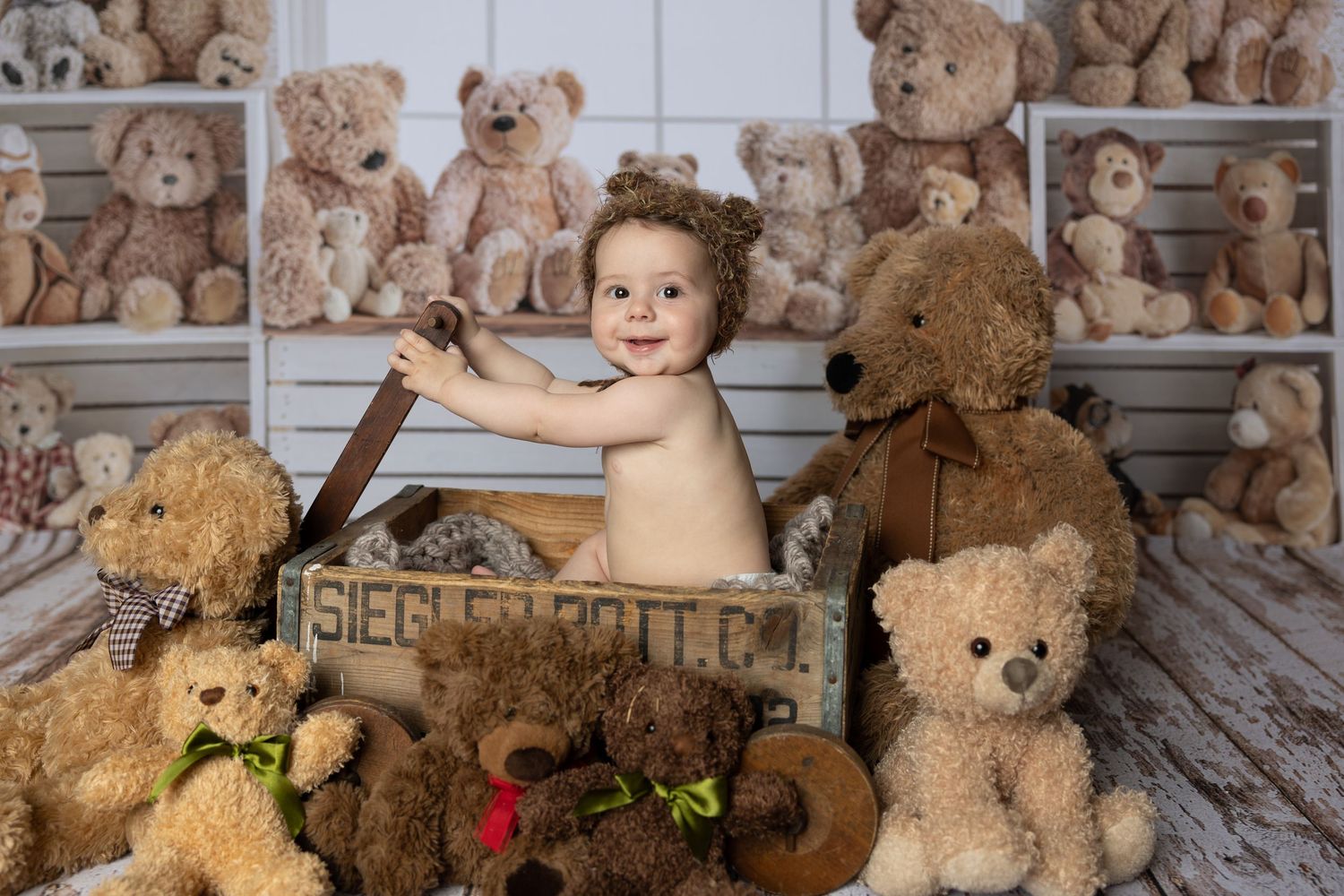 Smiling baby sitting in wooden crate surrounded by multiple brown teddy bears on white shelving backdrop.